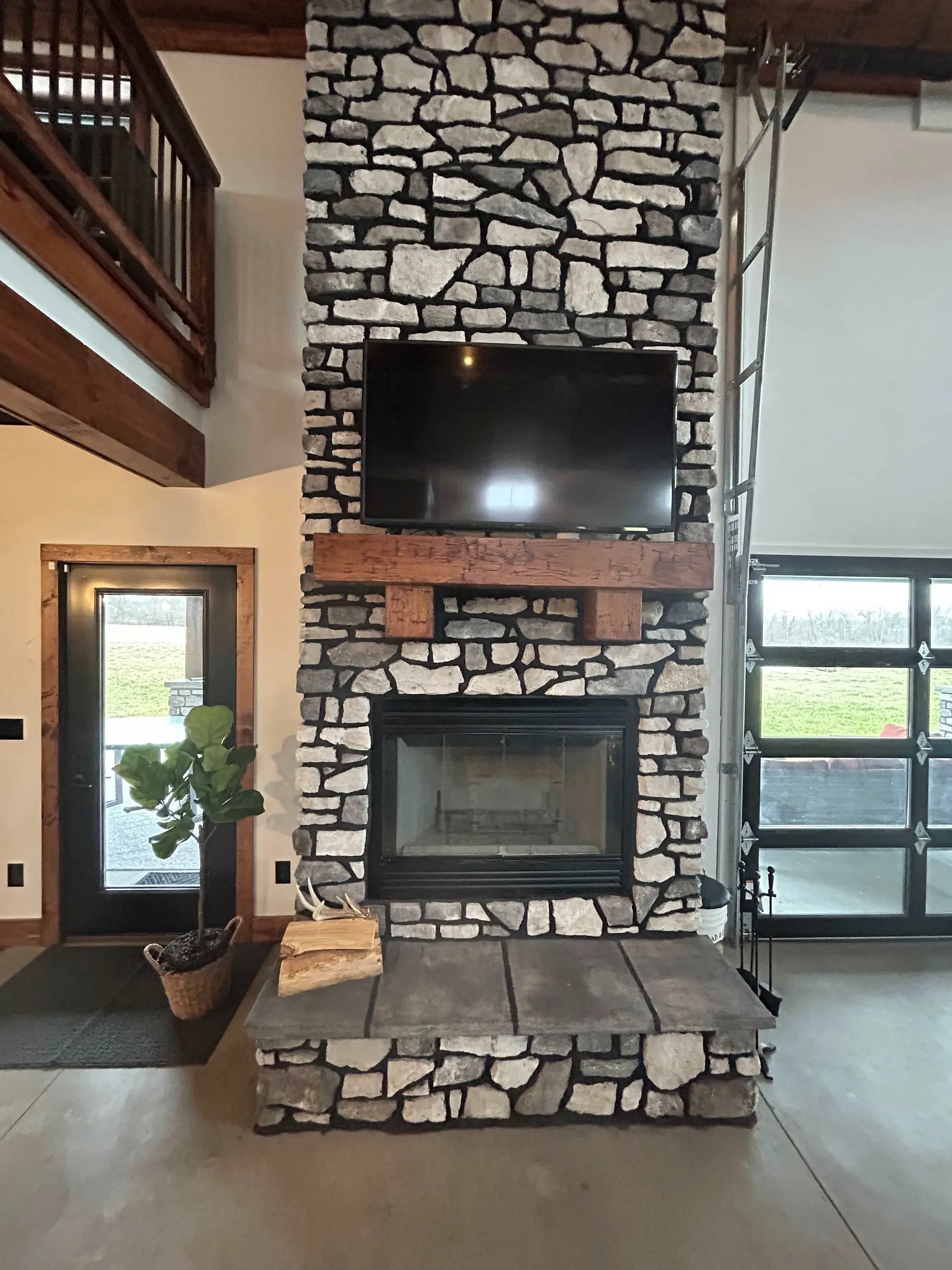 Rustic living room with stone fireplace, TV, wood mantle, and large windows.