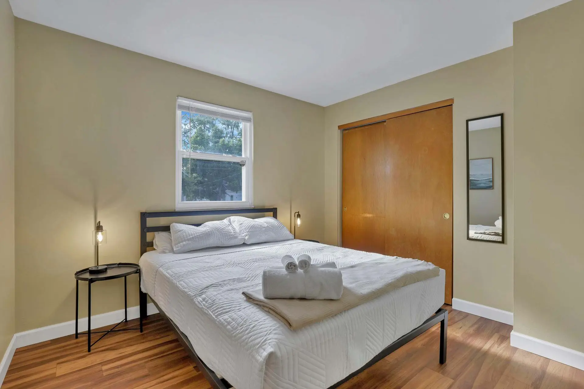 A neatly made bed in a room with tan walls and a wood floor.
