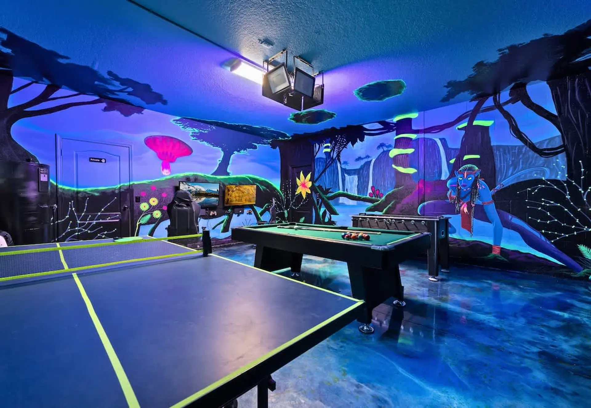 Room with ping pong table, pool table, and foosball table, decorated with neon murals.