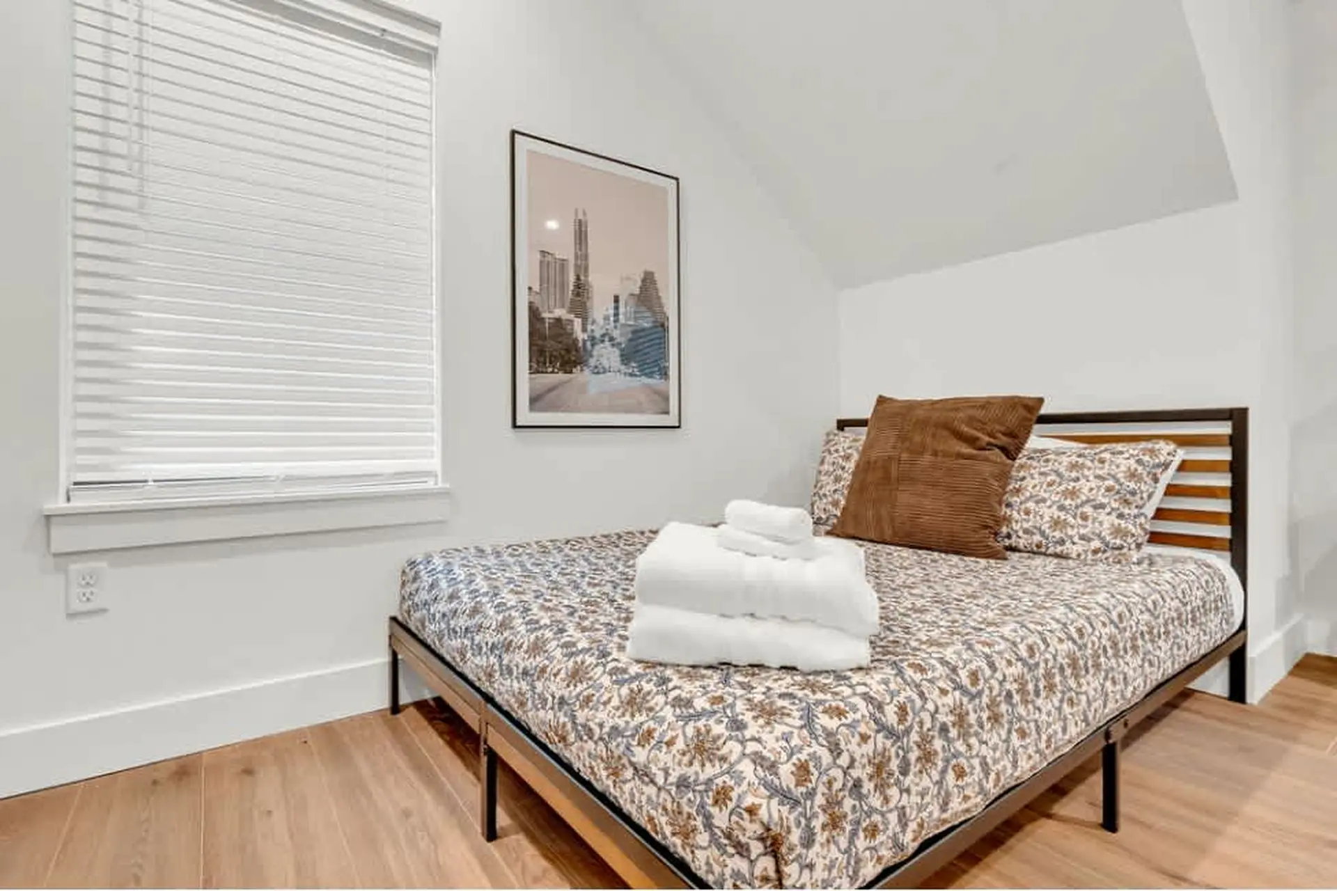 A bed with floral print bedding and stacked towels, next to a framed picture of a city skyline.