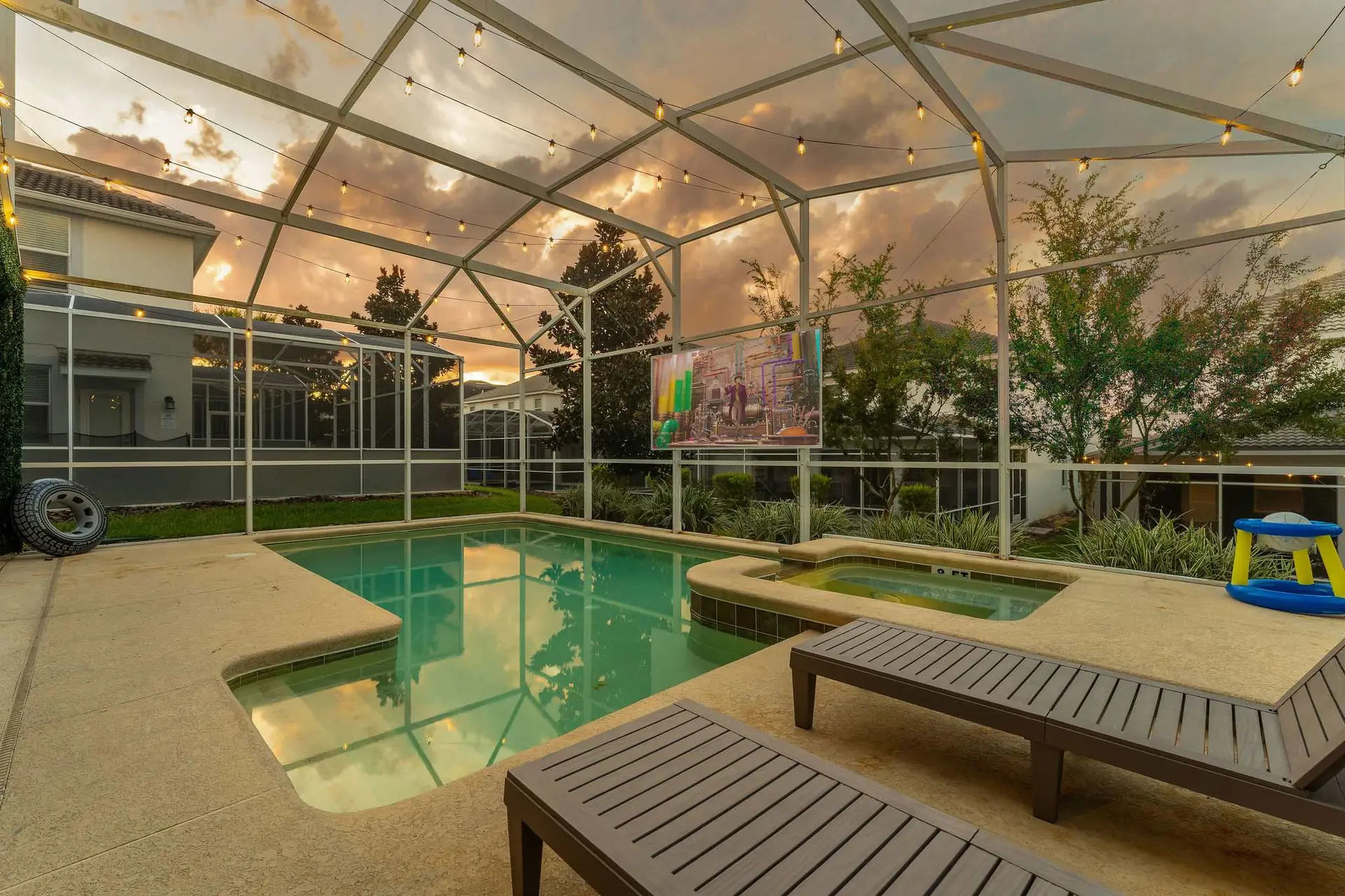 Pool and spa area with lounge chairs and a large outdoor TV under string lights at sunset.