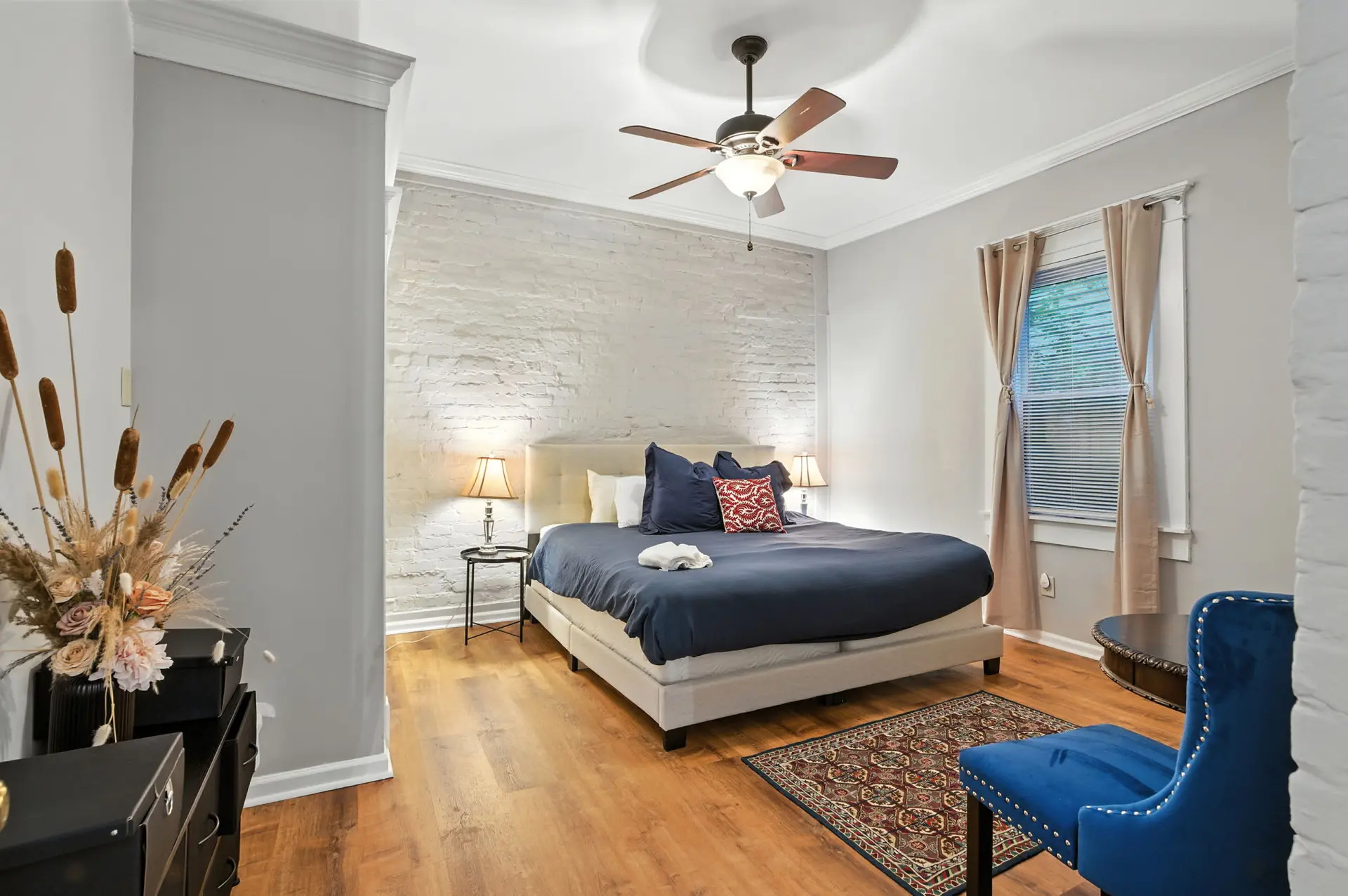 A bedroom with a white brick wall, a king-size bed with navy bedding, and a ceiling fan.