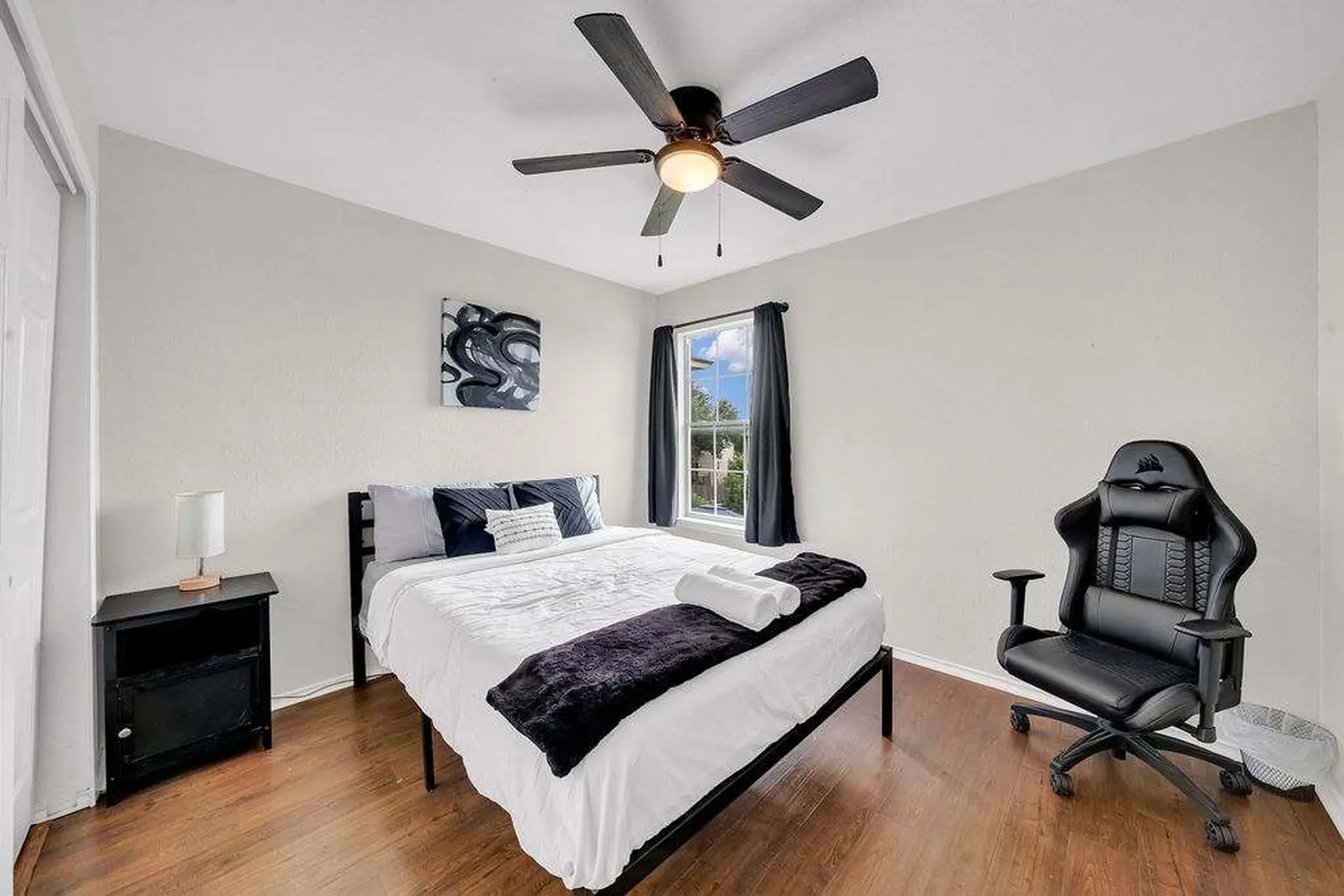 A black gaming chair sits on a wooden floor next to a bed and a nightstand.