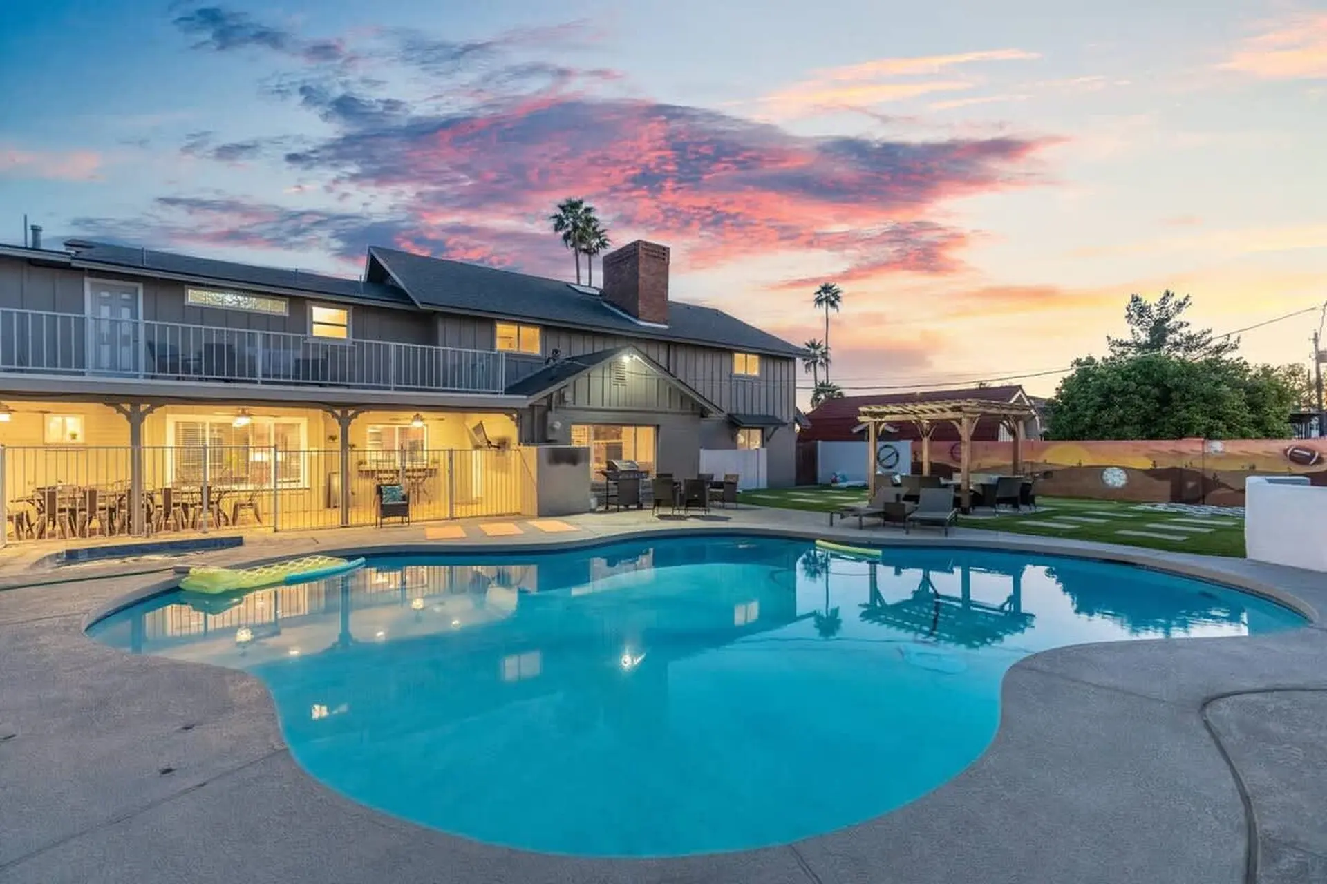 A large swimming pool in the foreground, with a modern house and a backyard patio.