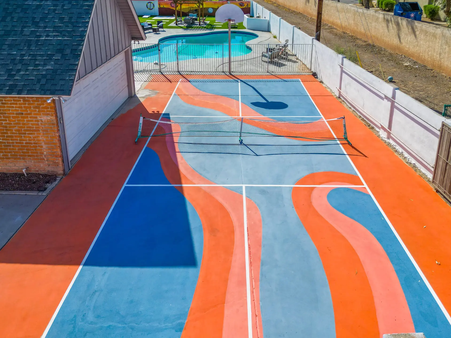 An aerial view of a blue and orange pickleball court with a swimming pool and basketball hoop in the