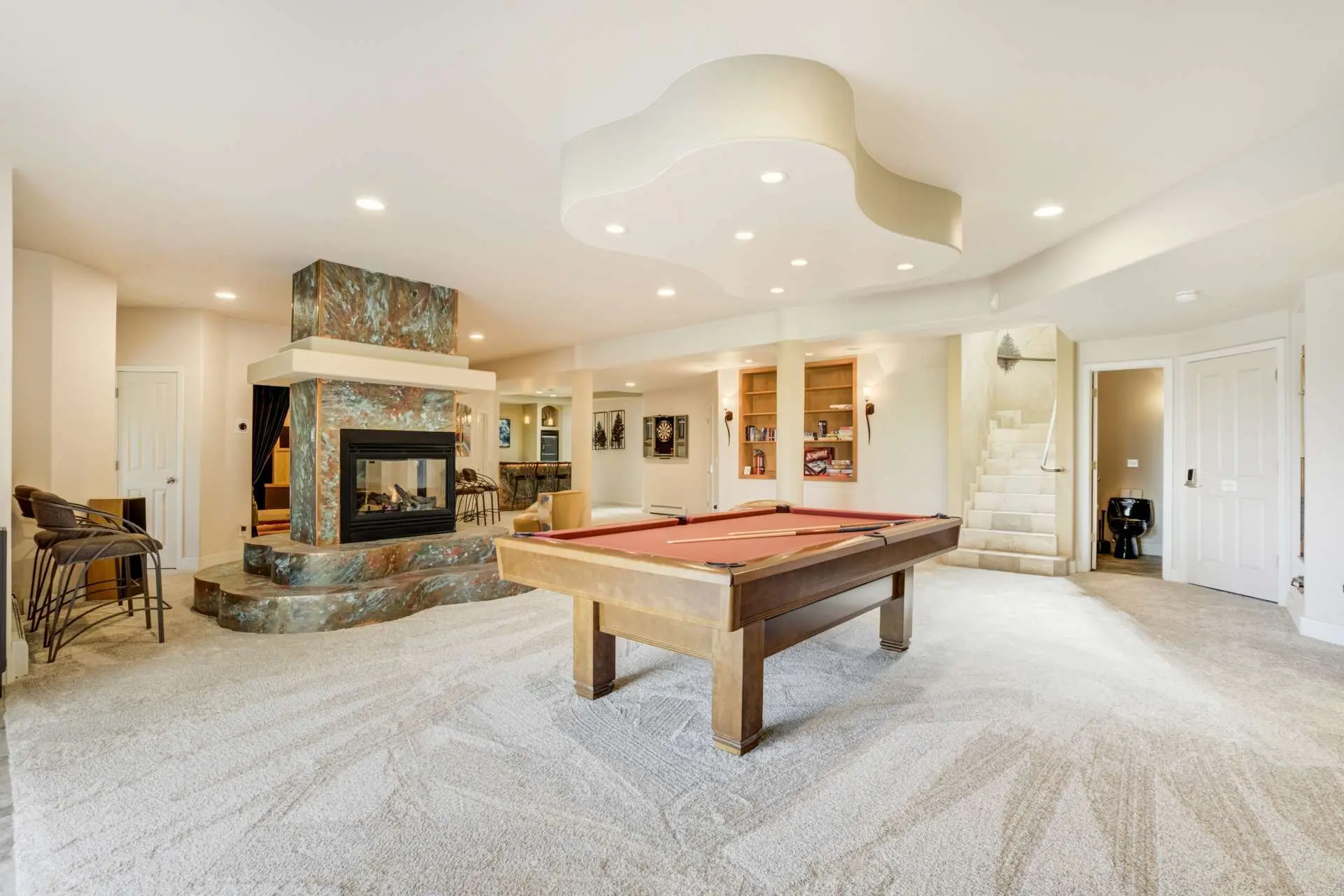 A basement recreation room with a pool table, fireplace, bar, and staircase.