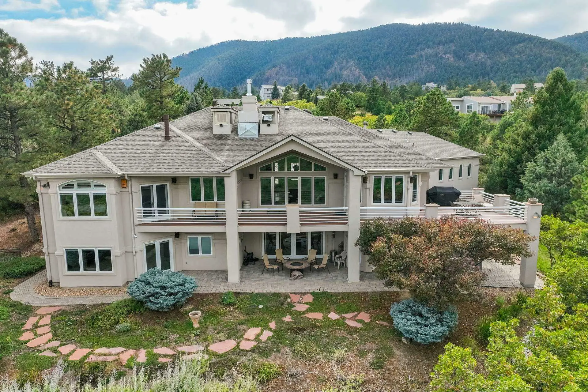 Aerial view of a large, modern home with a deck, patio, and fire pit, set against a mountain backdro