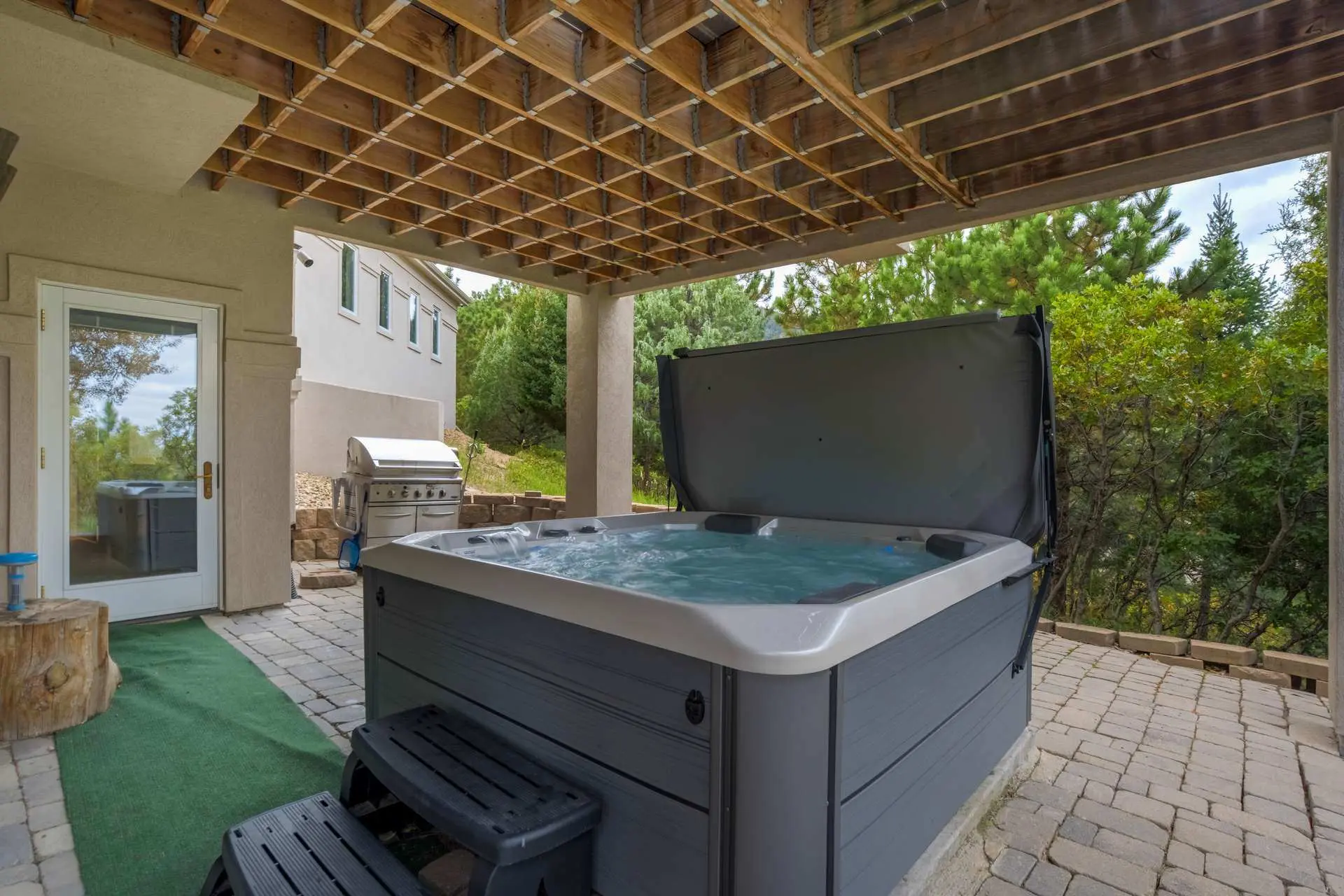 Hot tub and grill on paved patio under wooden deck with trees in background.