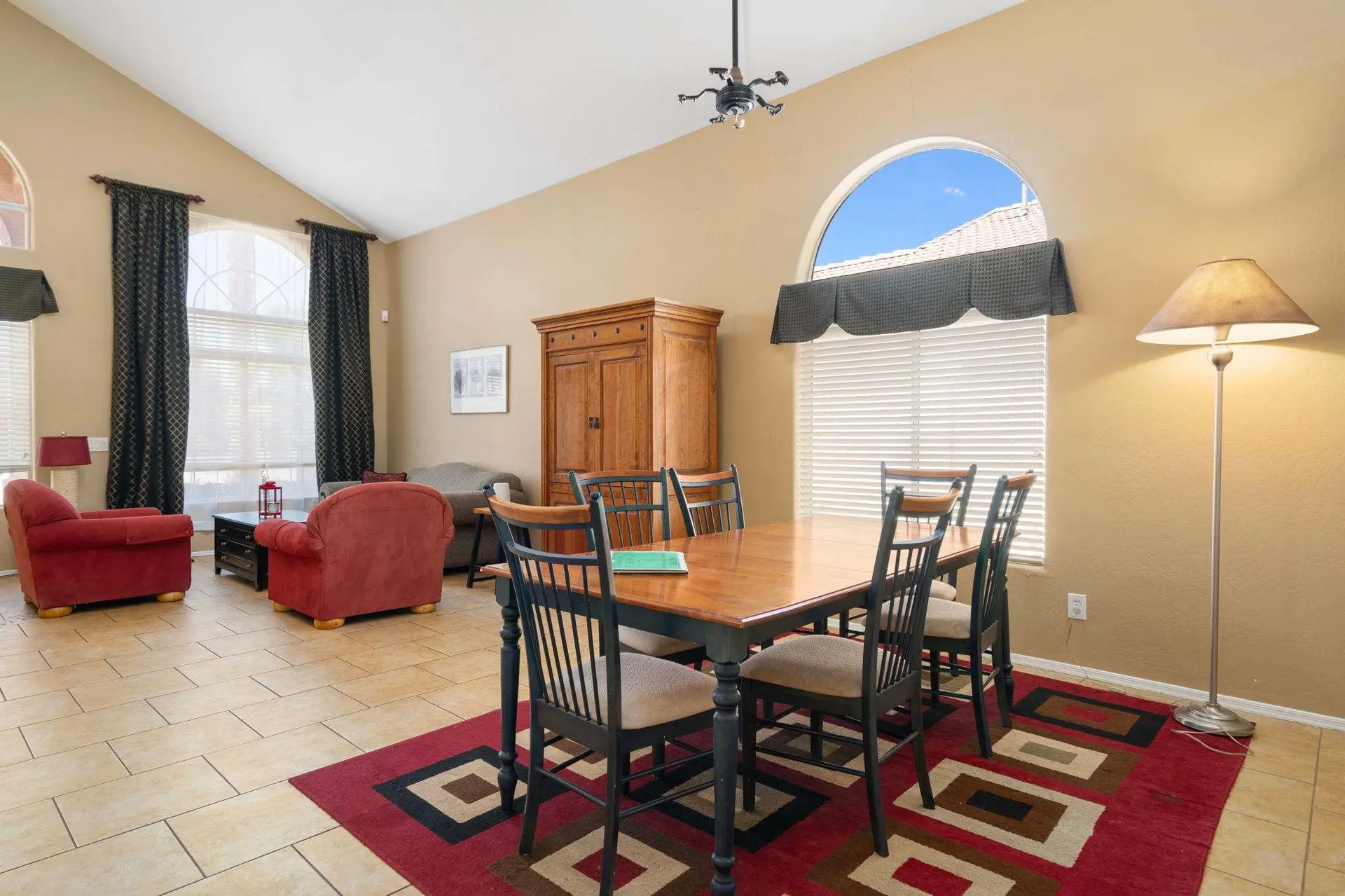 A dining room with a wooden table and chairs on a patterned rug, next to a large arched window.