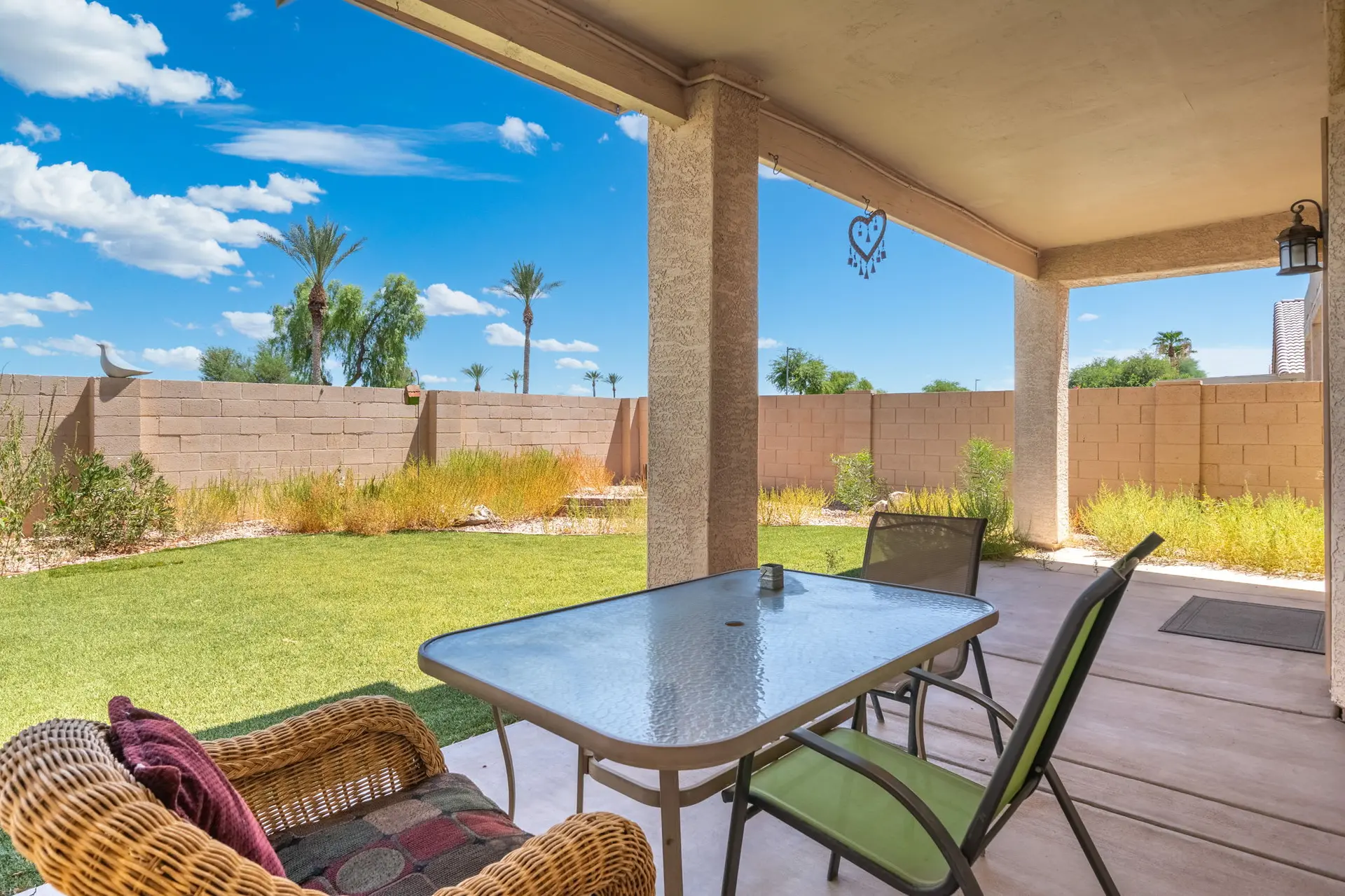 A patio with a glass-top table, chairs, and a wicker chair overlooks a green lawn and a brick wall.