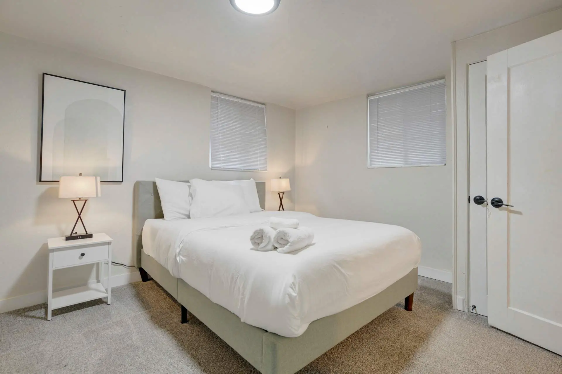 A bedroom with a bed, nightstand, framed art, and two windows.