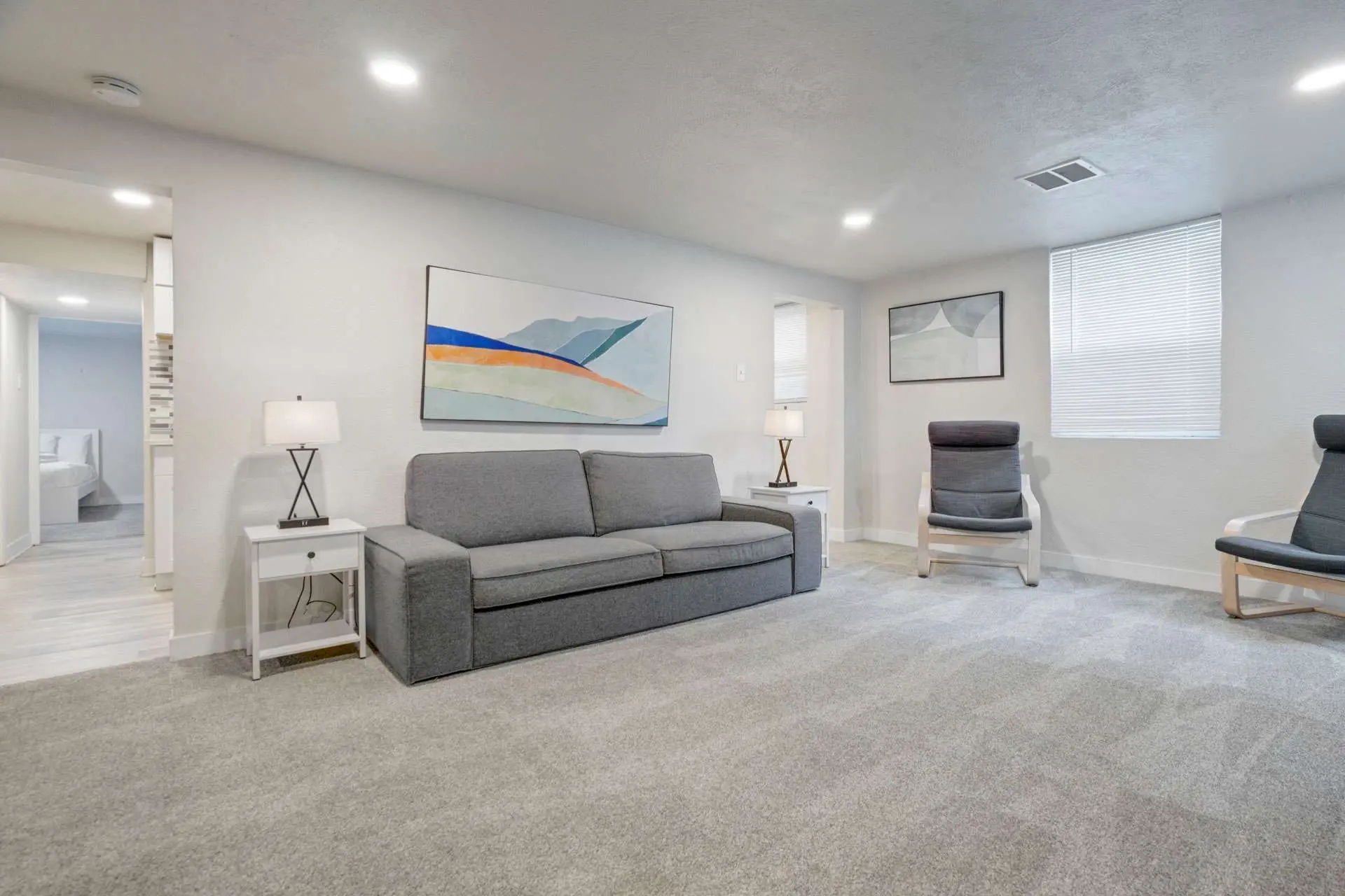 A living room with gray sofa, chairs, artwork, and a smoke detector on the ceiling.