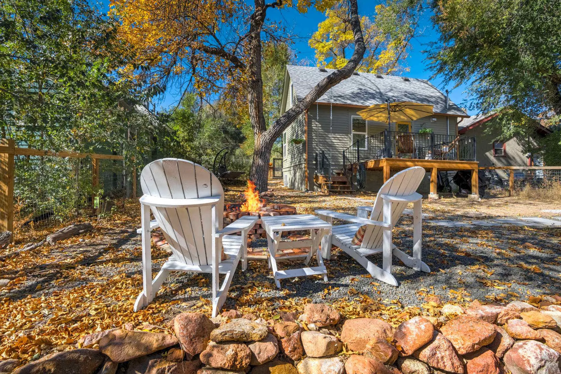 A cozy backyard scene with Adirondack chairs around a lit fire pit, surrounded by autumn leaves and 