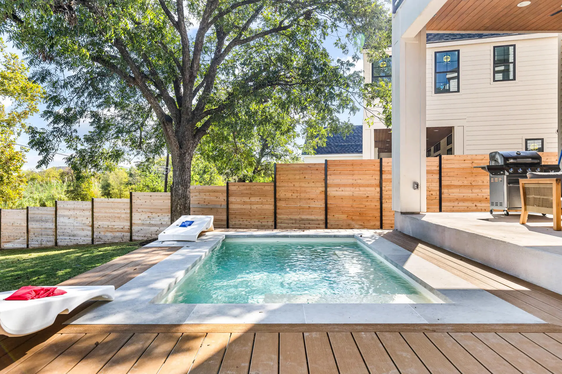 Modern backyard with a small pool, lounge chairs, grill, and wooden fence.