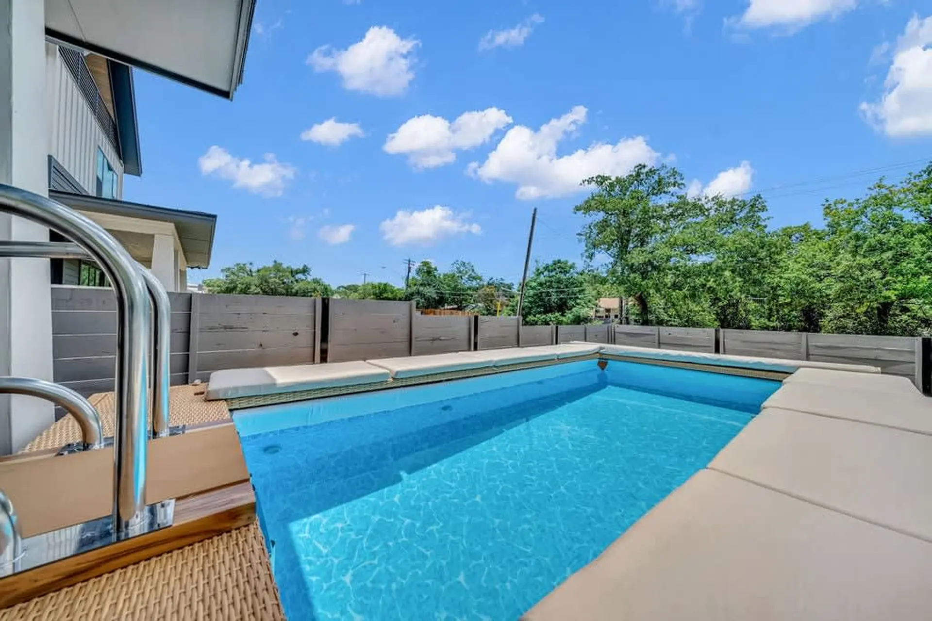 A crystal blue swimming pool with a modern design, surrounded by a wooden deck and a contemporary fe