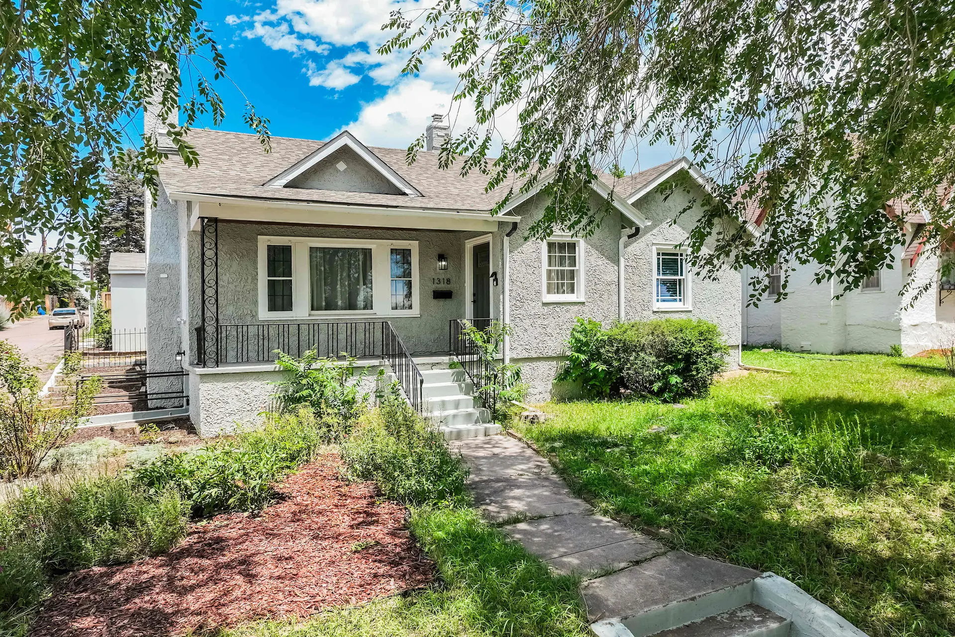 A grey, stucco house with white trim and windows, front porch with railings, and a sidewalk.
