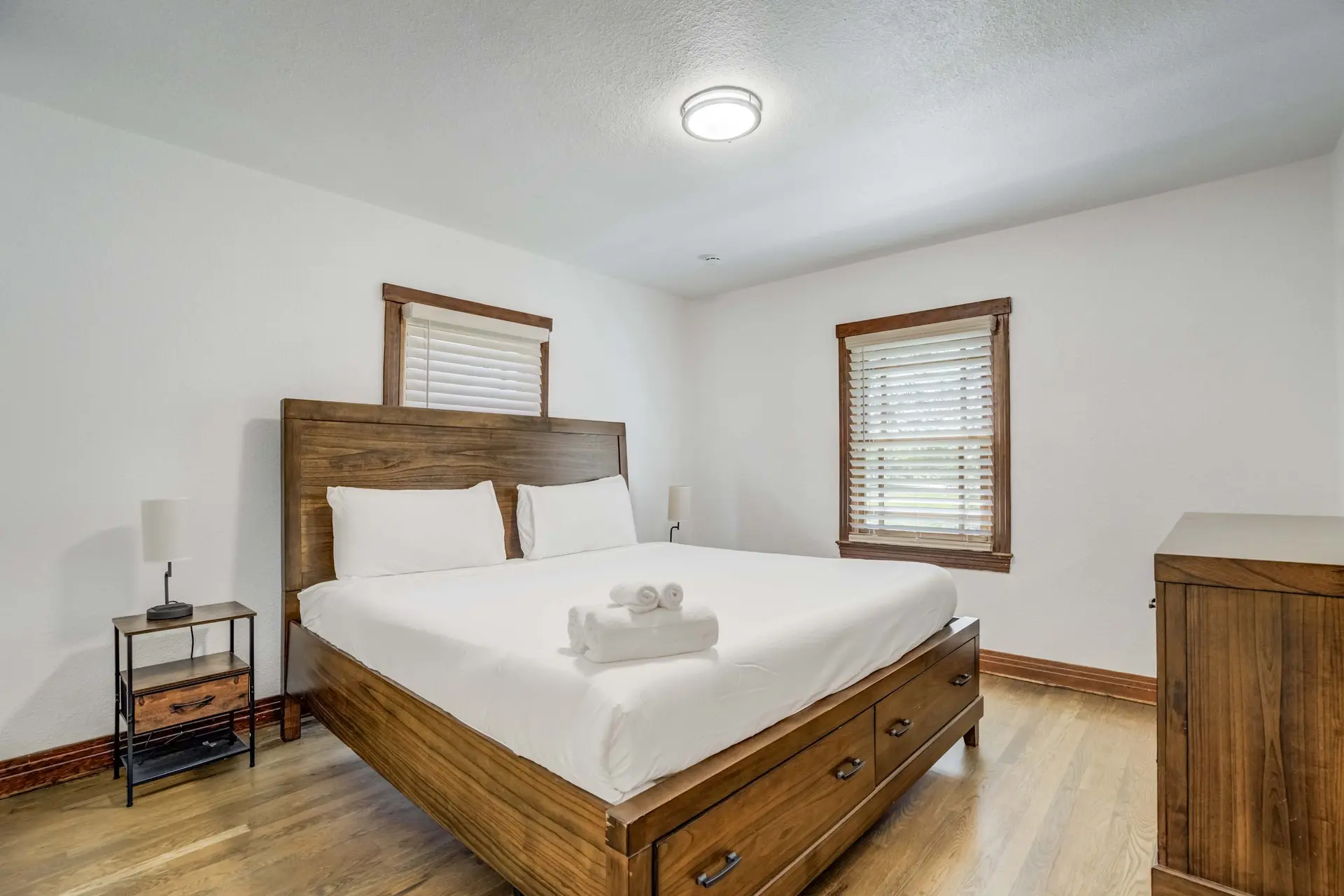 A bedroom with a wooden bed, nightstand, dresser, and two windows.