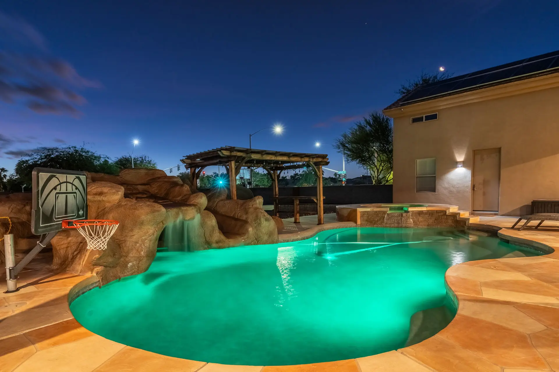 Luxury backyard with glowing pool, waterfall, hot tub, and basketball hoop at twilight.