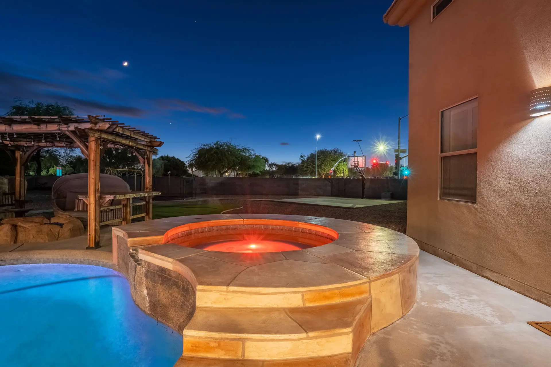 Luxury backyard with a pool, hot tub, basketball court, and pergola at dusk.