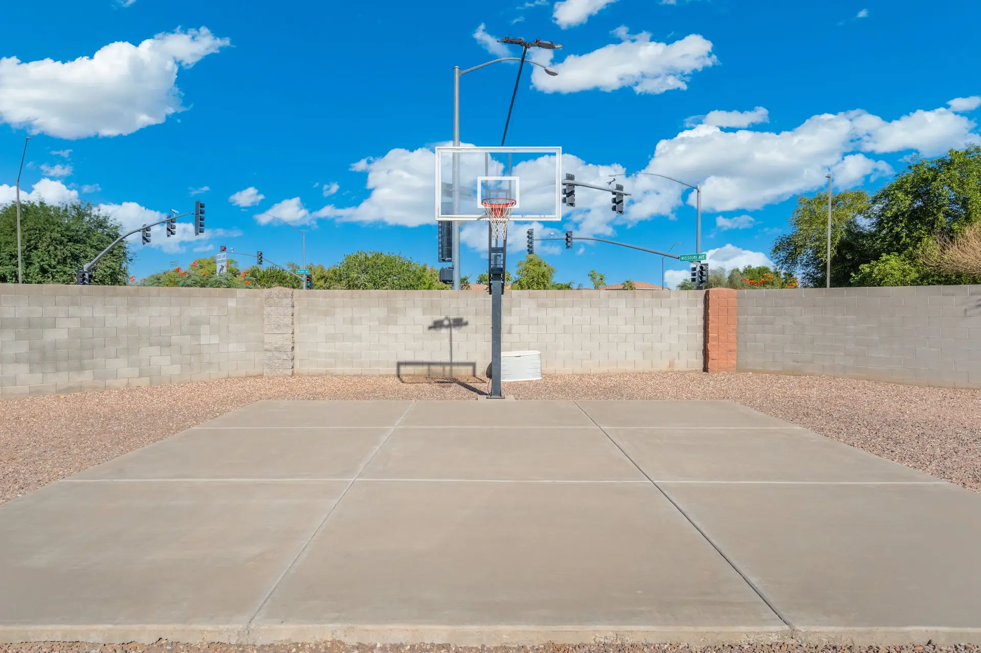 Outdoor basketball court with hoop, concrete court, gravel, and a cinder block wall under a blue sky