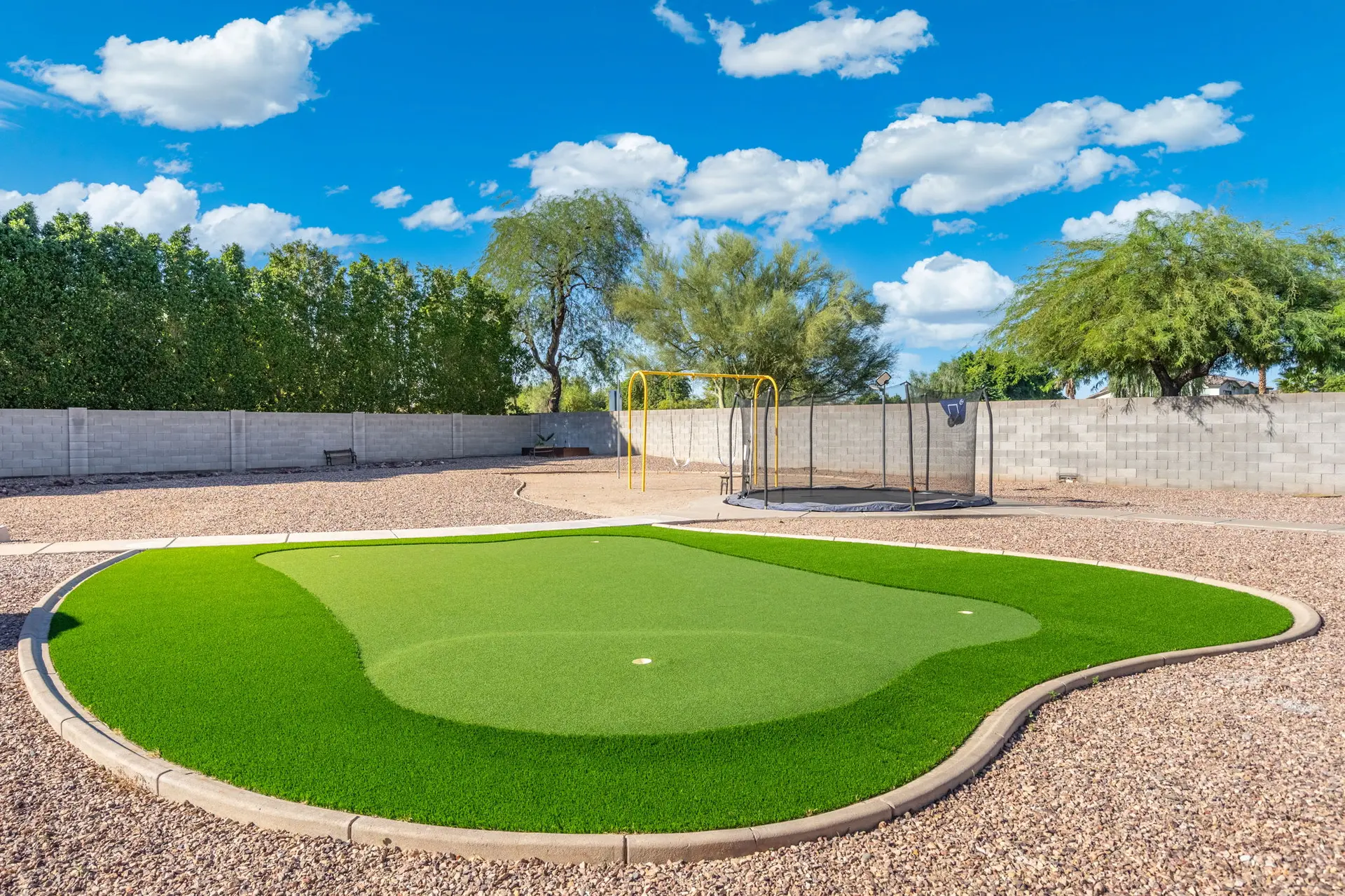 Backyard with a putting green, trampoline, and swing set under a blue sky.
