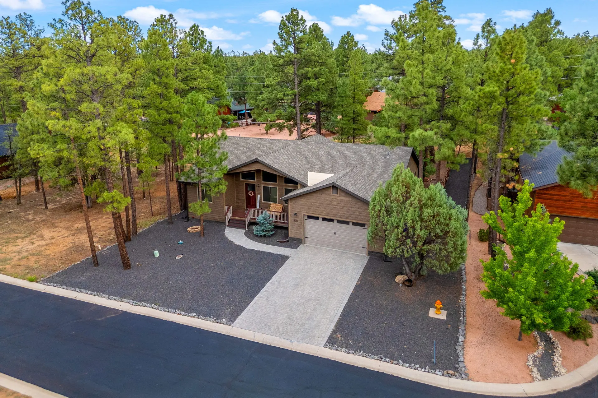 An aerial view of a modern brown house nestled amongst tall pine trees in a suburban neighborhood.