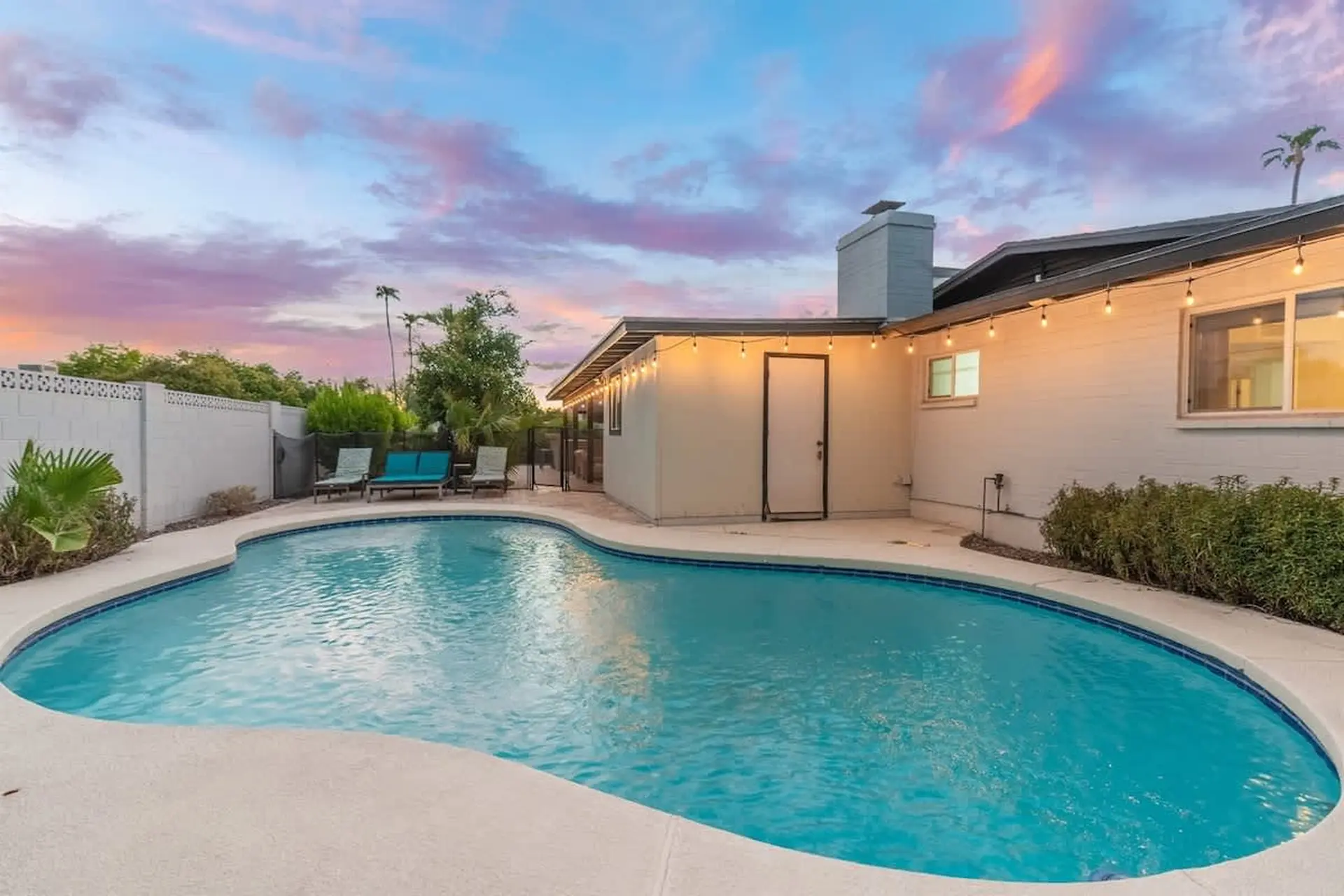 A backyard pool with lounge chairs, a white brick house, and a colorful sunset sky.