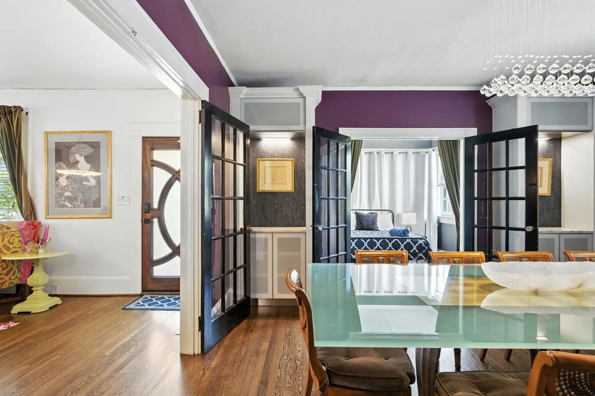 A dining room with a glass table and chairs, a chandelier, and a bedroom in the background.