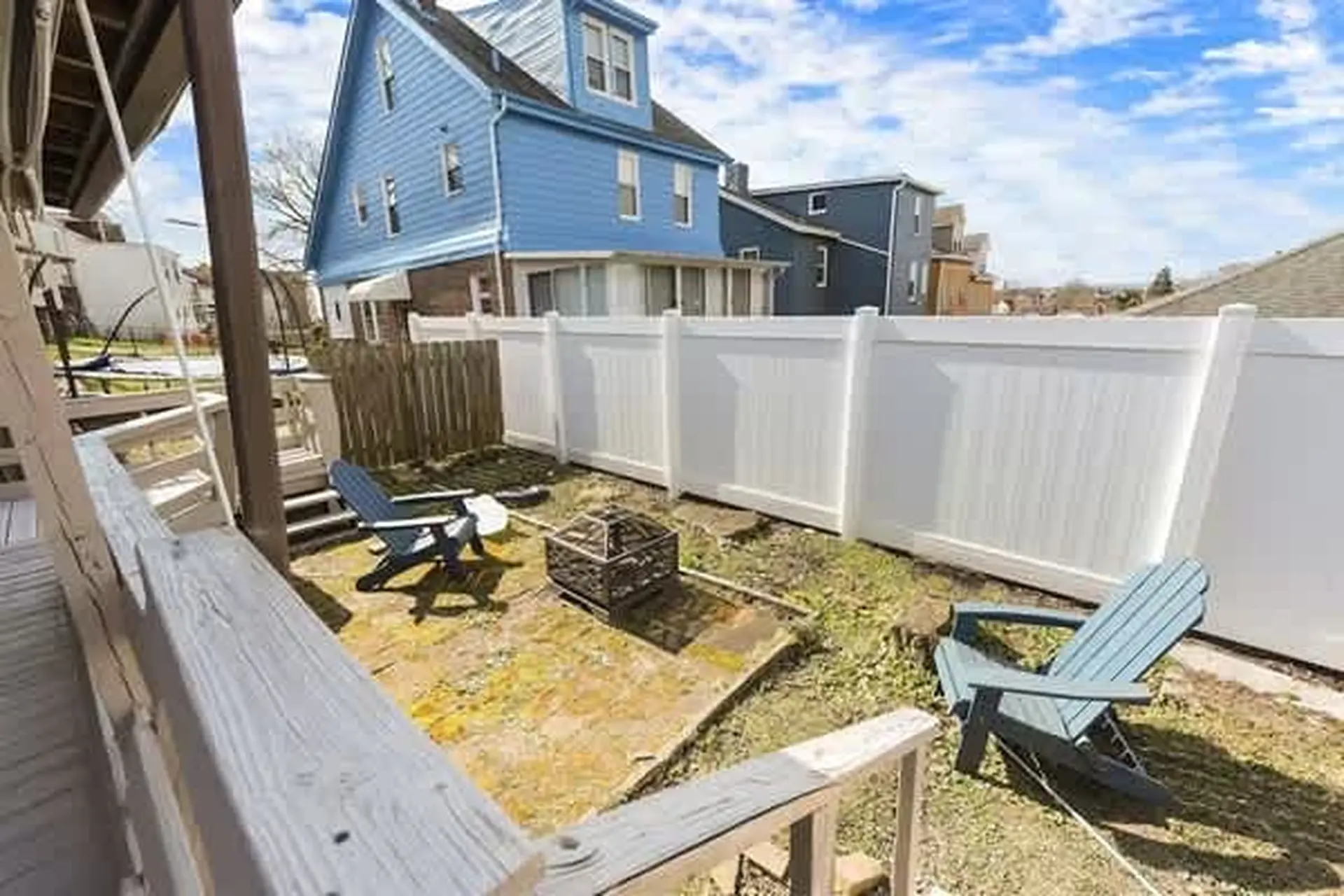 A backyard scene with a blue house, white vinyl fence, and two Adirondack chairs.