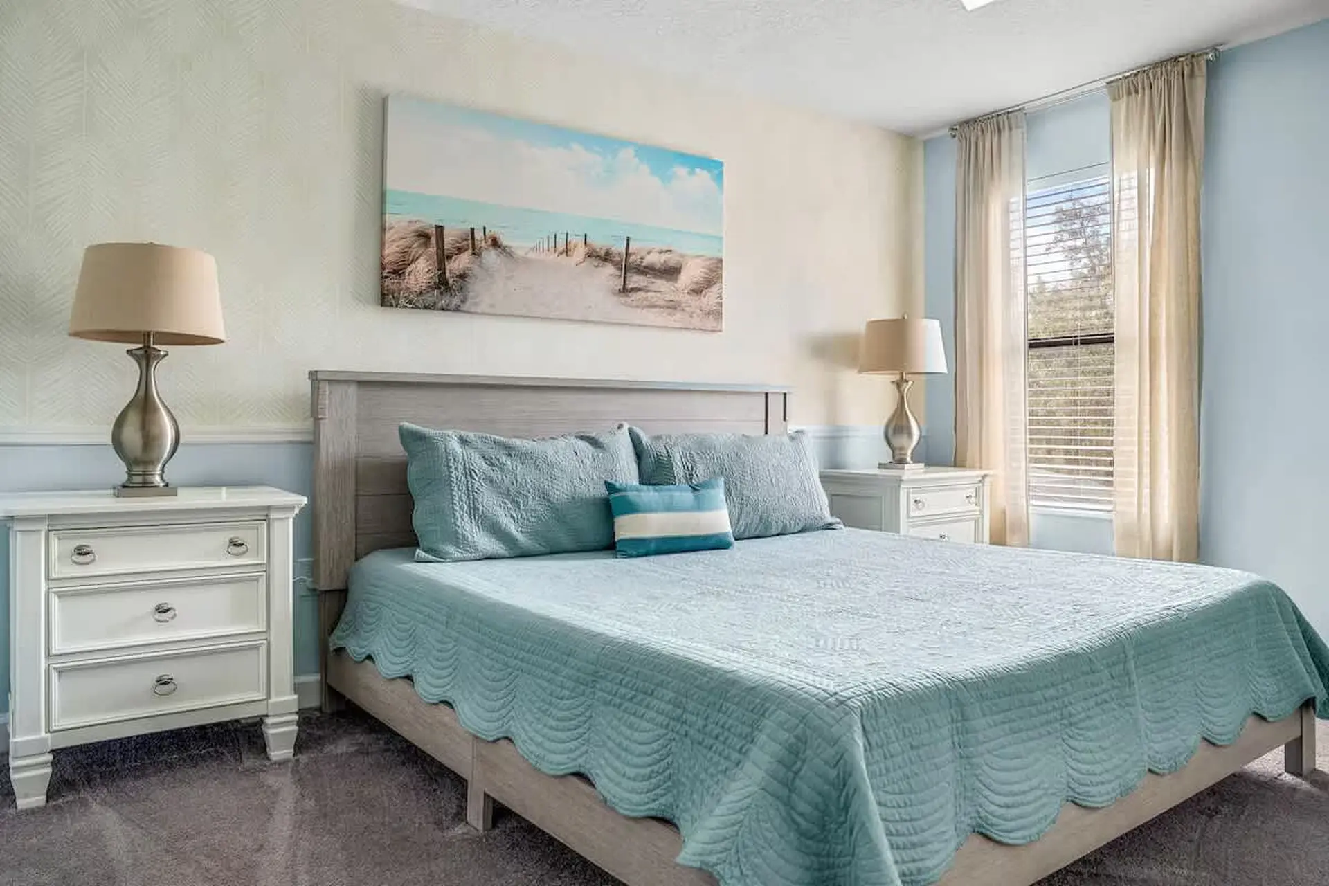 A coastal bedroom features a bed with blue bedding, a large beach scene painting, and a window with 