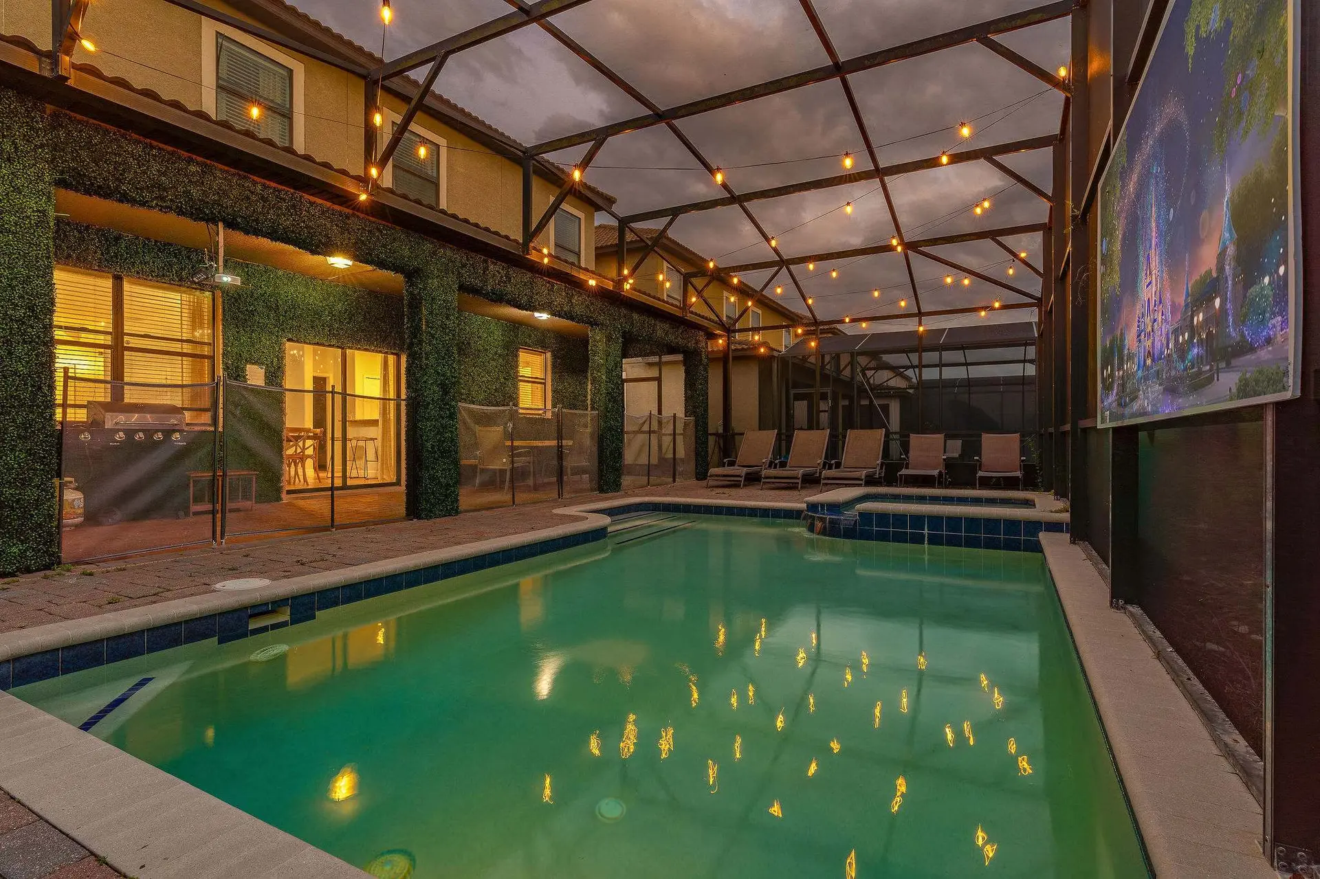 A screen-enclosed pool and patio area with string lights, lounge chairs, and a large outdoor screen.