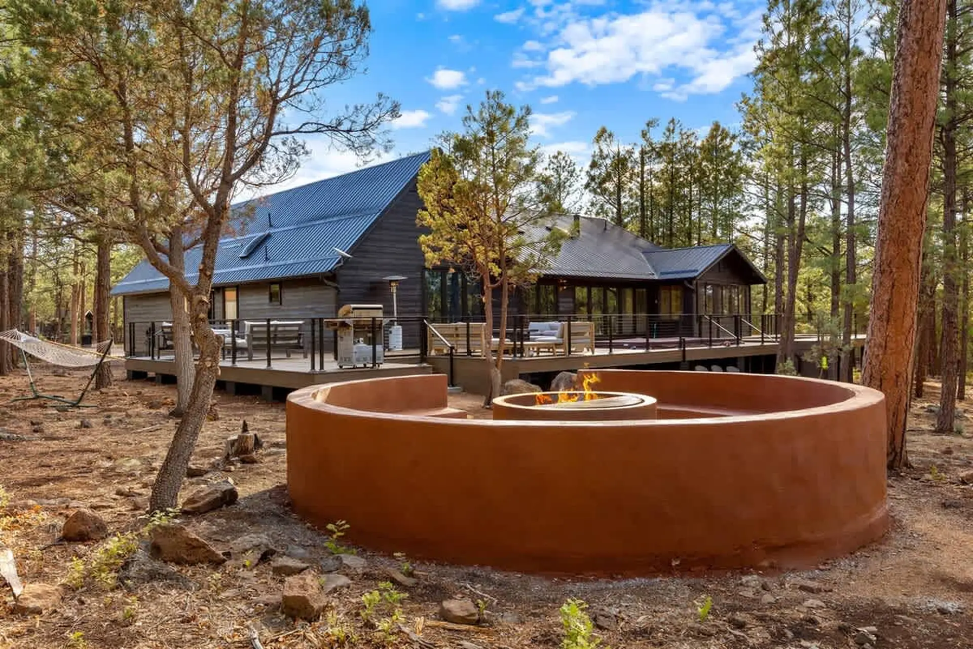 Modern cabin in a forest with a large deck and a circular fire pit.