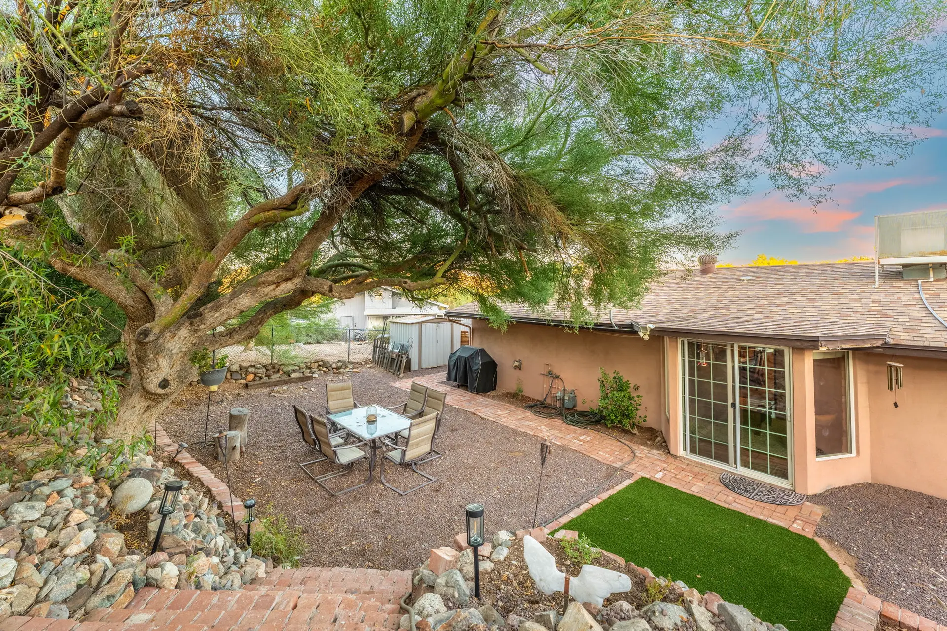 Backyard patio with a large tree, dining set, grill, and artificial turf.