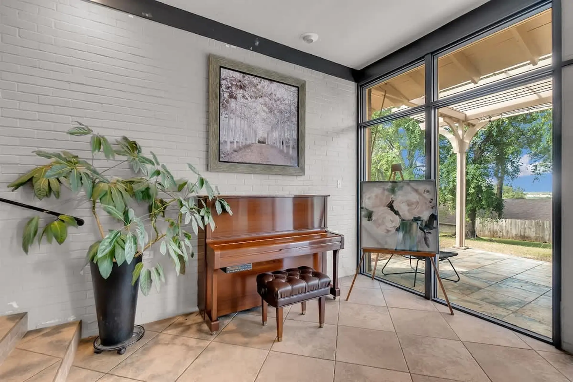 Piano and artwork in a room with a large window overlooking a patio and garden.
