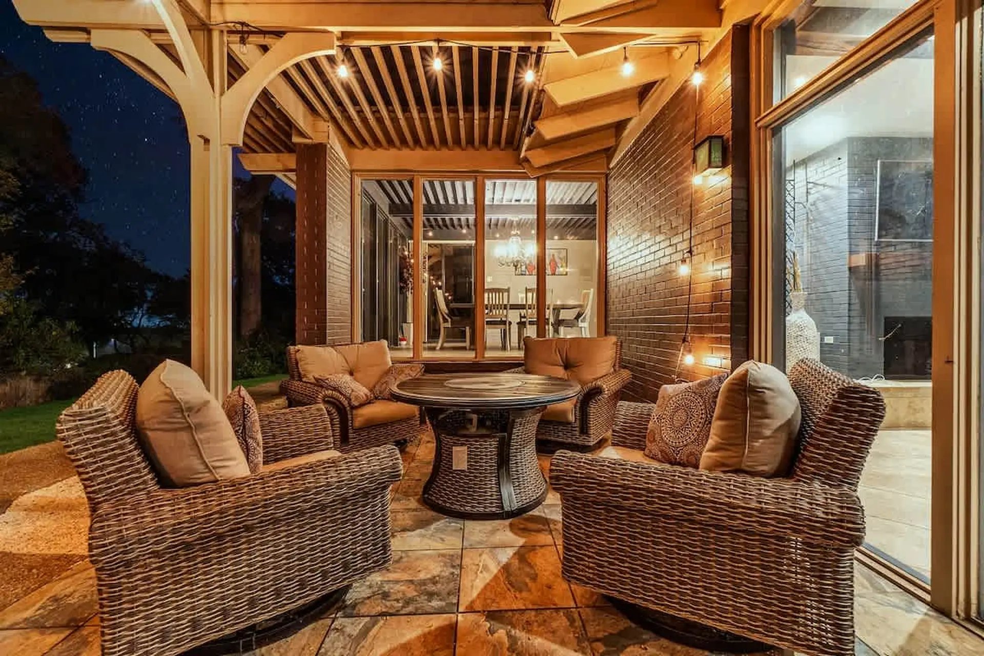 An outdoor patio with wicker furniture and a round table, illuminated by string lights.