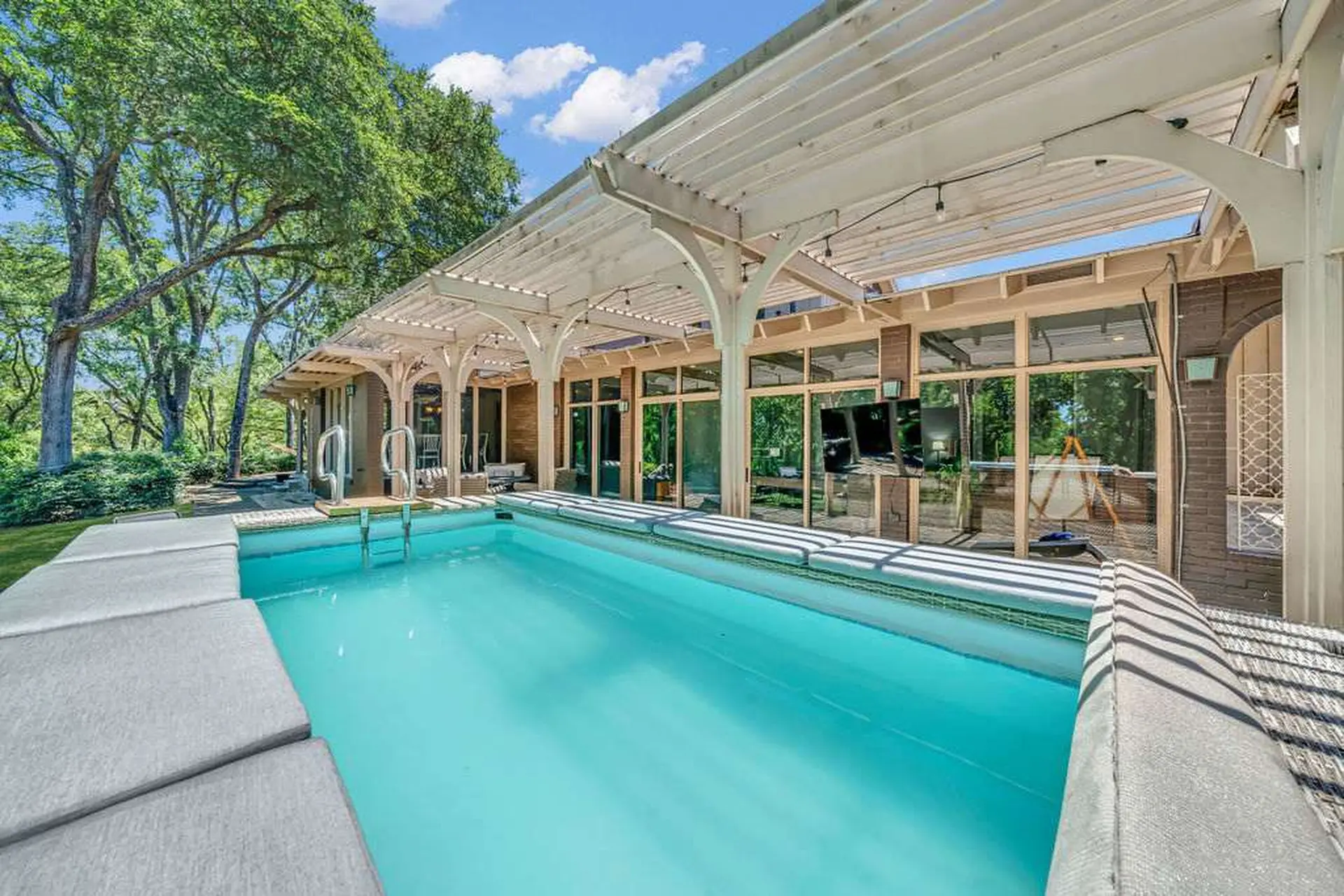 A bright blue swimming pool with a white pergola-covered patio and lush green trees.