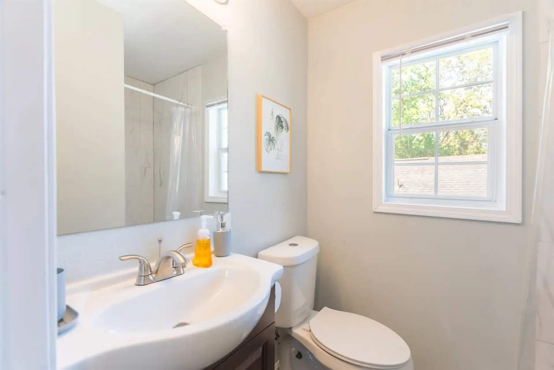 A modern bathroom with a white sink and toilet, a large mirror, and a window with a view of trees.