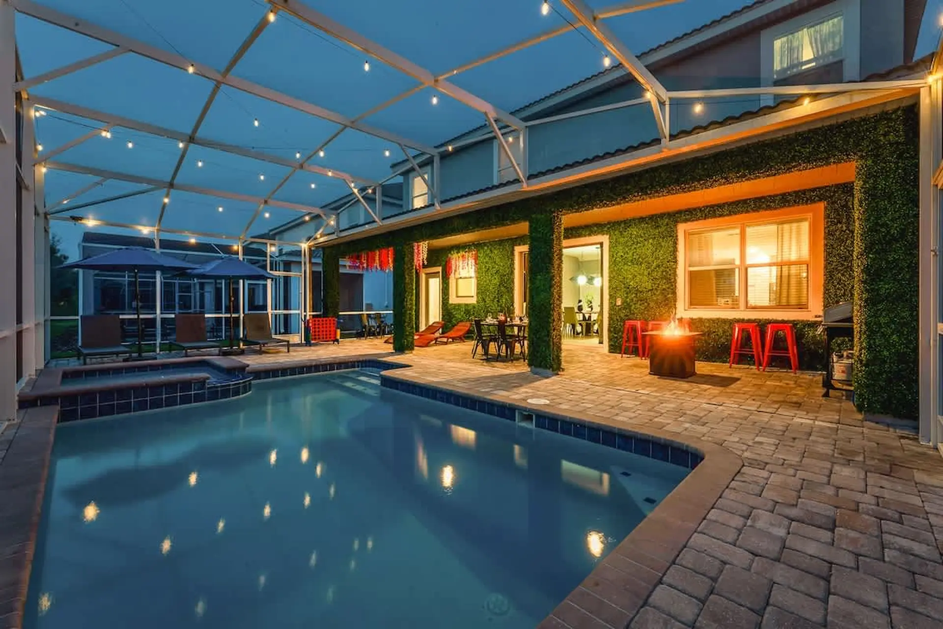A backyard pool and patio area at night, lit by string lights and a fire pit.