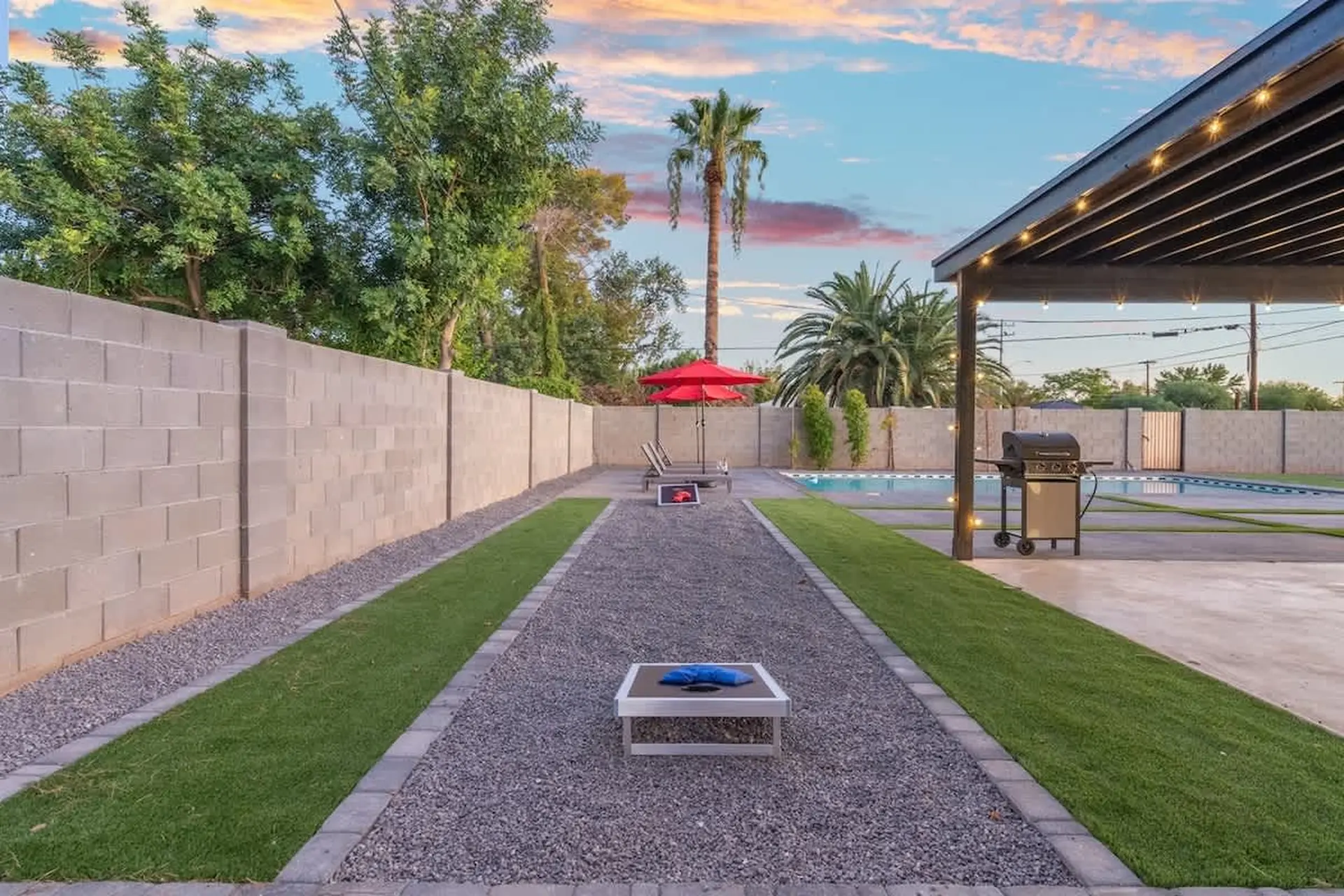 Backyard with pool, grill, cornhole game, and red umbrellas.