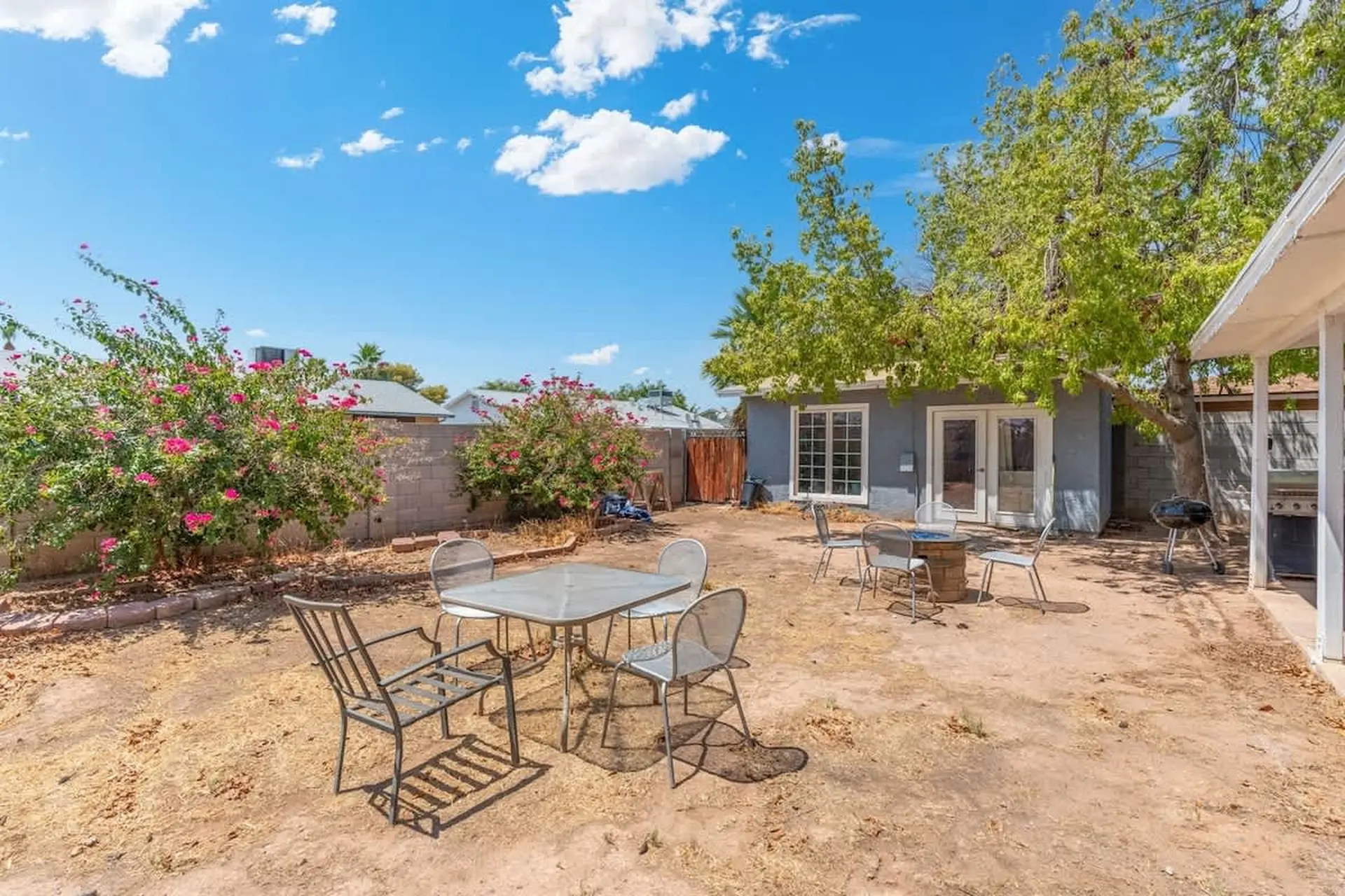 A backyard patio with a table, chairs, and a grill under a blue sky.