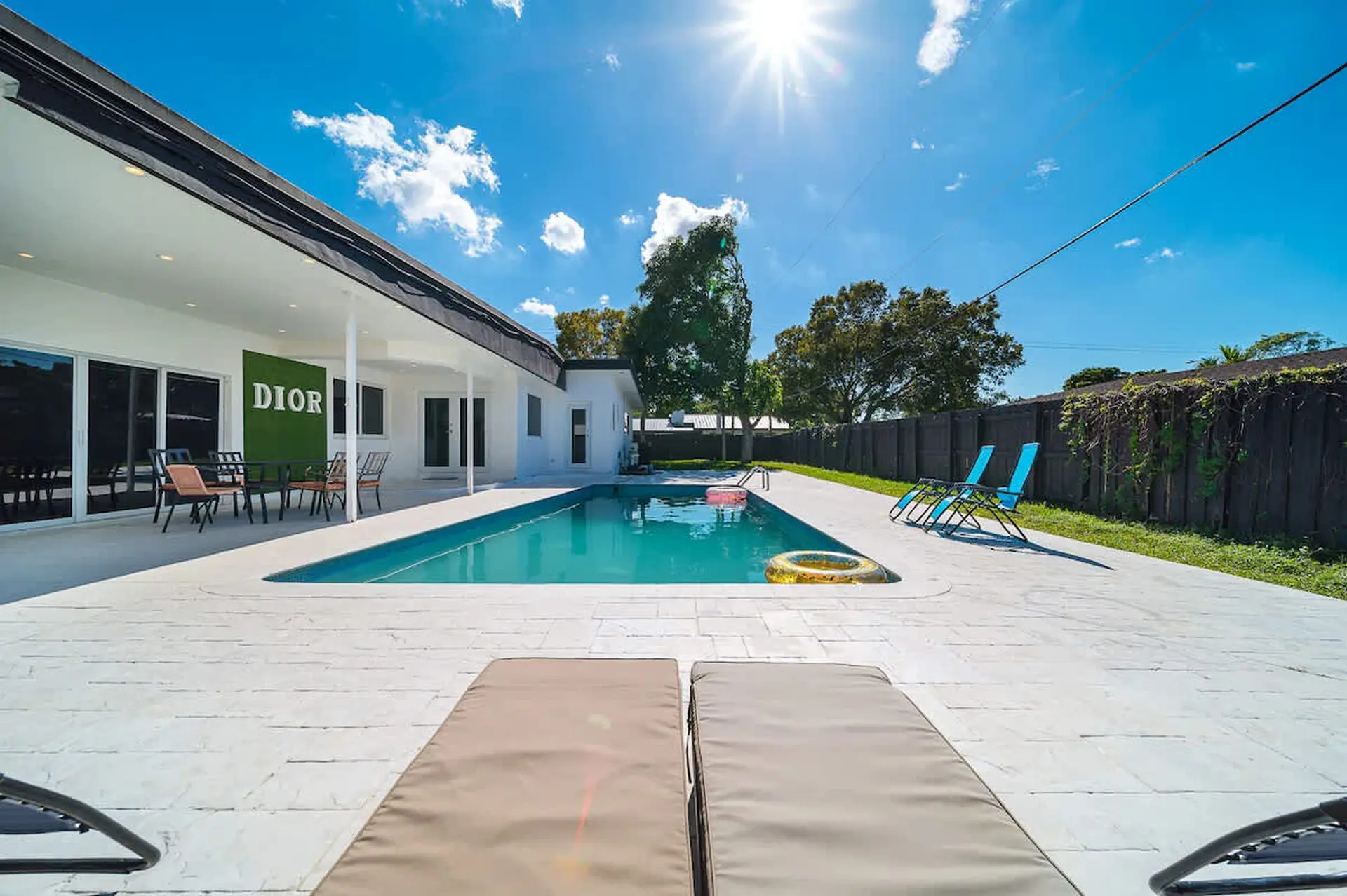 A sunny pool deck with lounge chairs, a pool with inflatables, and a house with a "DIOR" sign.