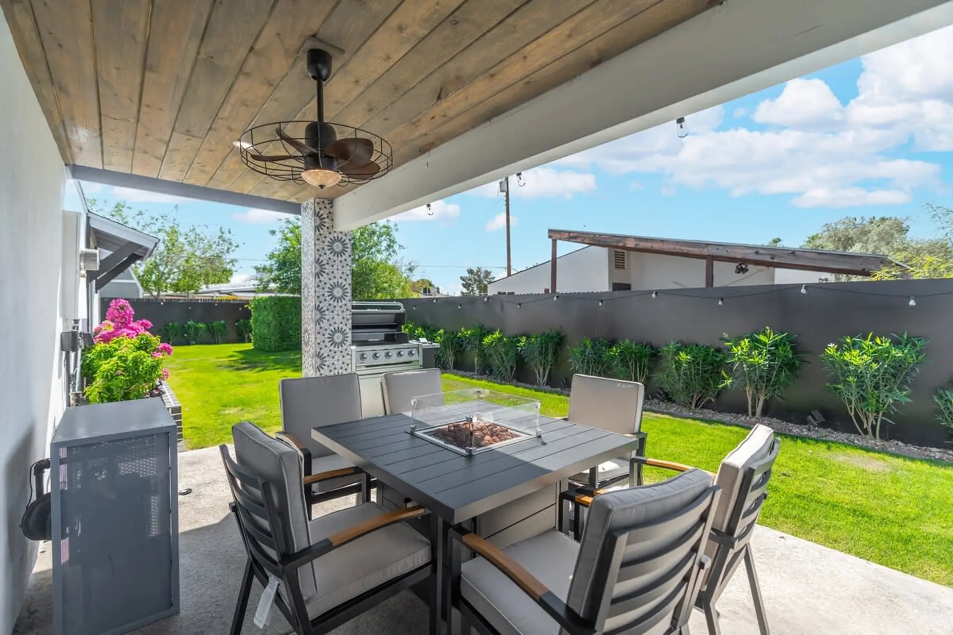 Outdoor patio dining set with a gas grill, covered by a wooden ceiling with a fan.