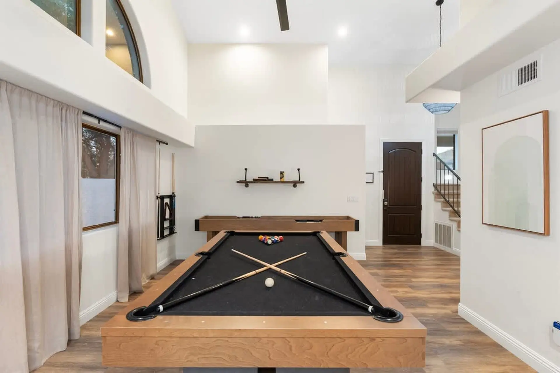 A pool table with cues and balls in a well-lit room with a staircase and a tall window.