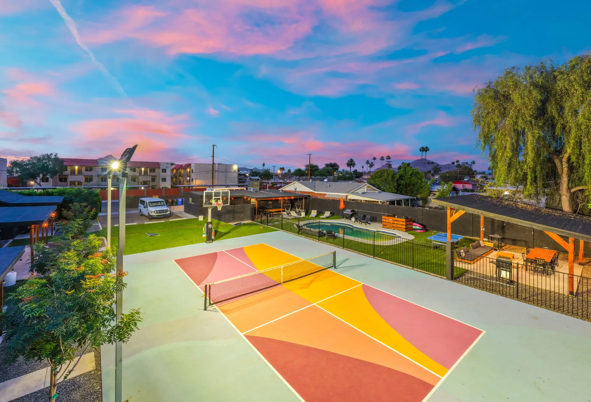 Colorful pickleball court at dusk with a pool, basketball hoop, and covered patio.