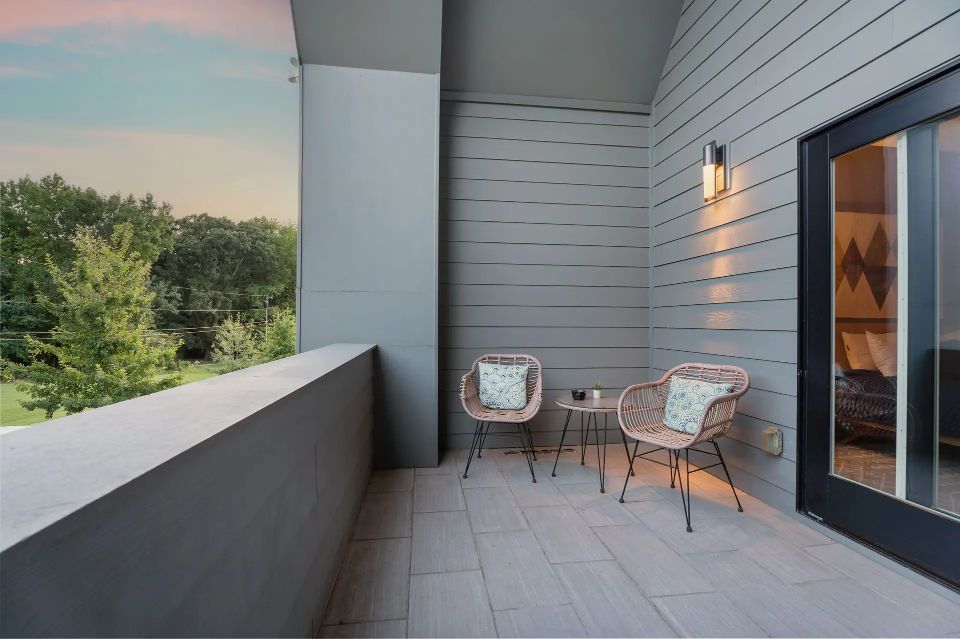 A balcony with two rattan chairs and a small table overlooks a tree-lined landscape at dusk.