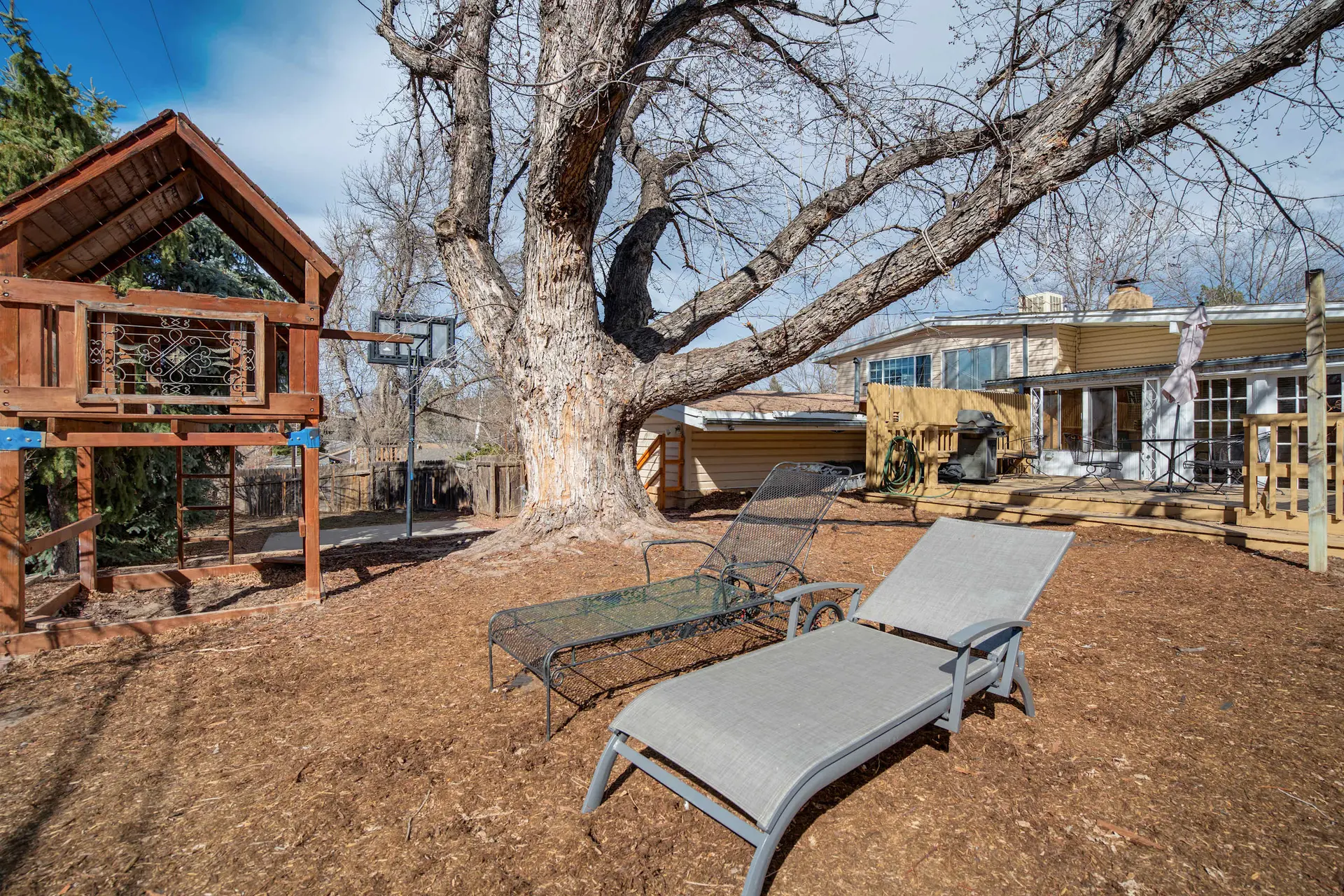 A backyard scene with a large tree, a wooden playhouse, two lounge chairs, and a deck.
