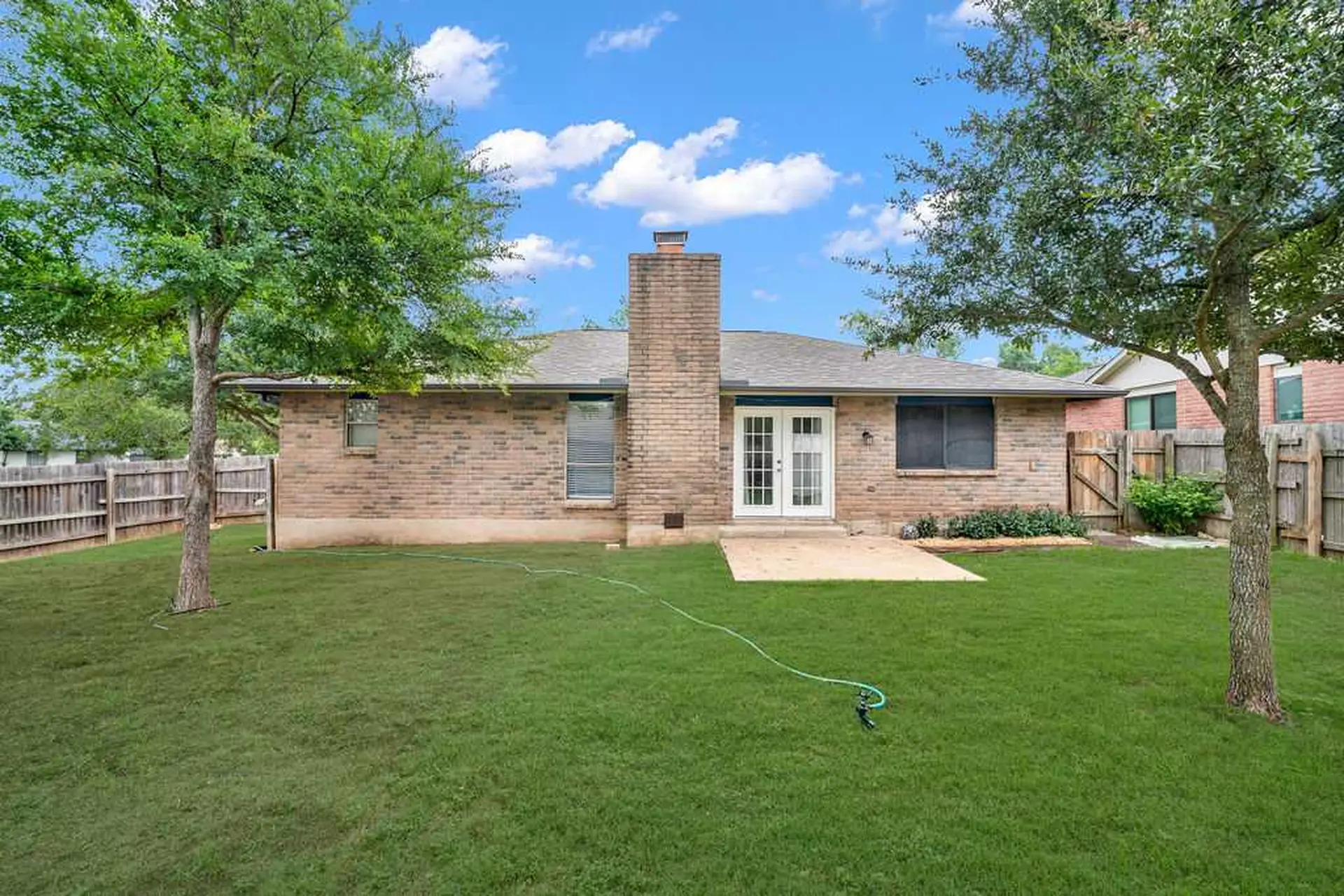 A backyard with a brick house, lawn, trees, and a fence.