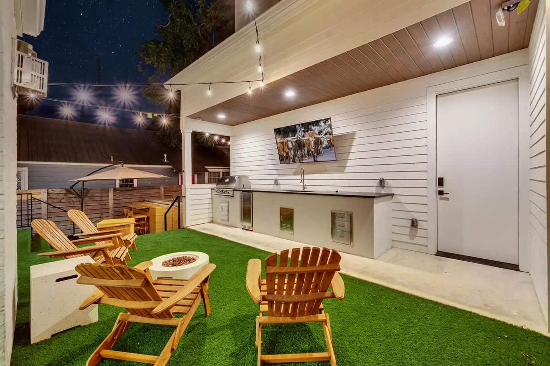 An outdoor patio with a grill, sink, TV, chairs, and a fire pit under a starry night sky.