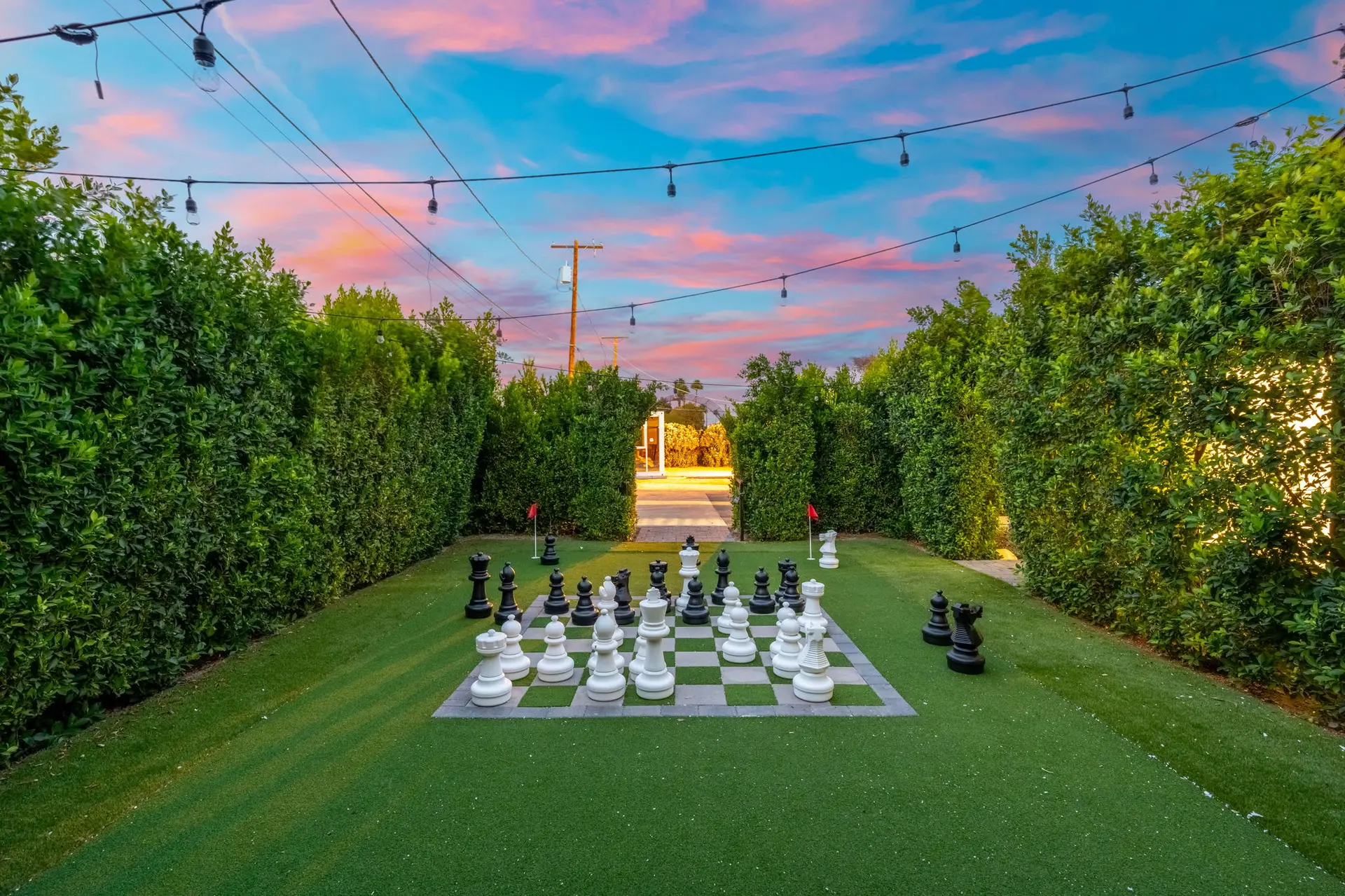 Giant chess set on artificial grass lawn surrounded by hedges with a colorful sunset sky.