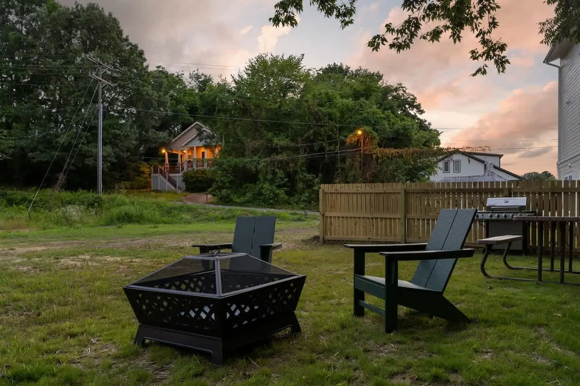 A backyard fire pit and chairs are set up for a gathering, with a grill and picnic table nearby.